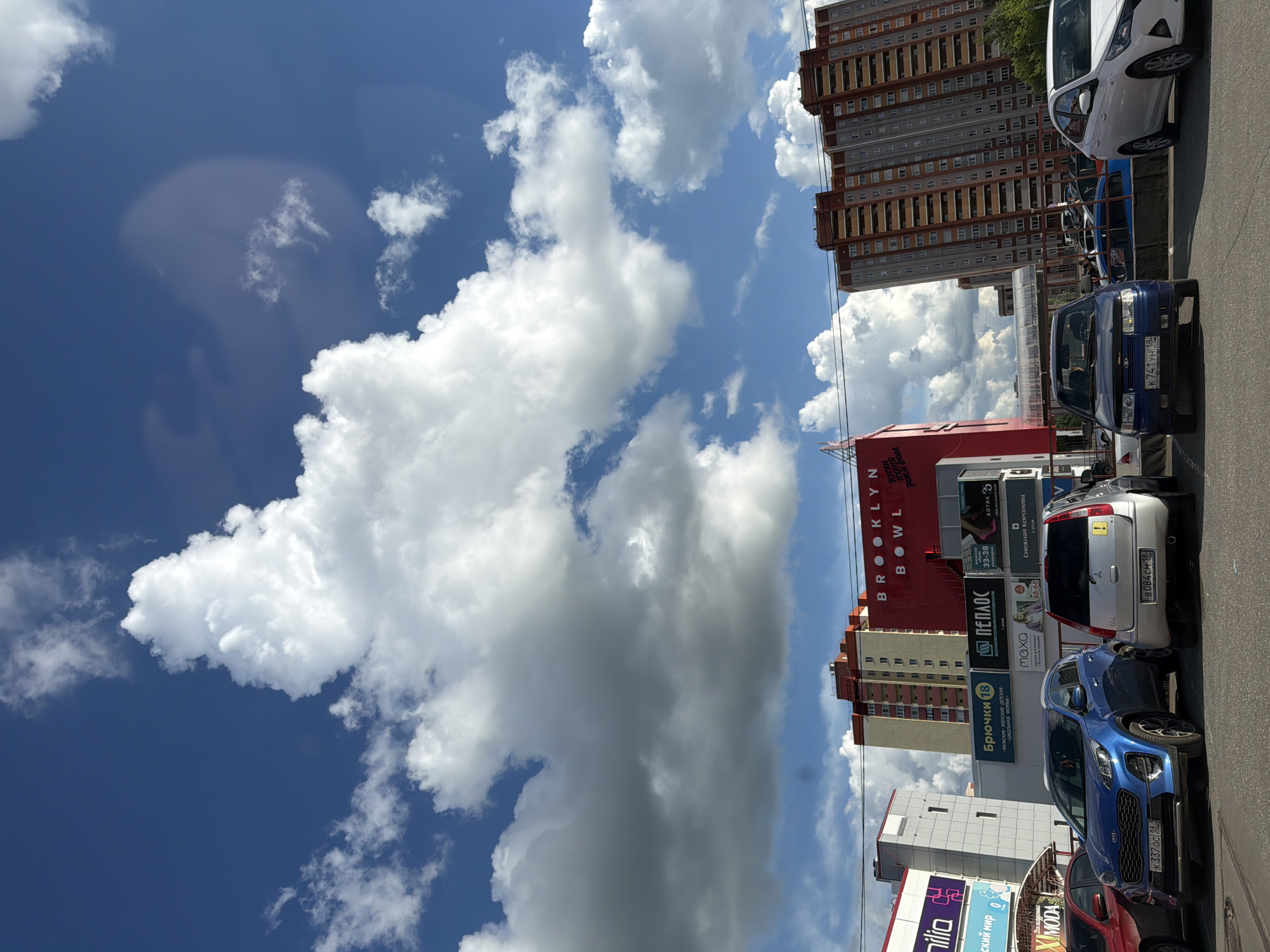 The image depicts a view of the sky with fluffy white clouds partially obscuring a bright blue background. In the lower portion of the frame, there is a building complex with commercial signage and parked cars. A red building with the text "BROOKLYN BOWL" is visible, along with advertisements for clothing stores and other businesses. The cars are parked along the street, adjacent to the buildings. Based on the signage, the image is likely taken in Izhevsk, Russia, specifically near the Brooklyn Bowl entertainment center and adjacent commercial establishments.