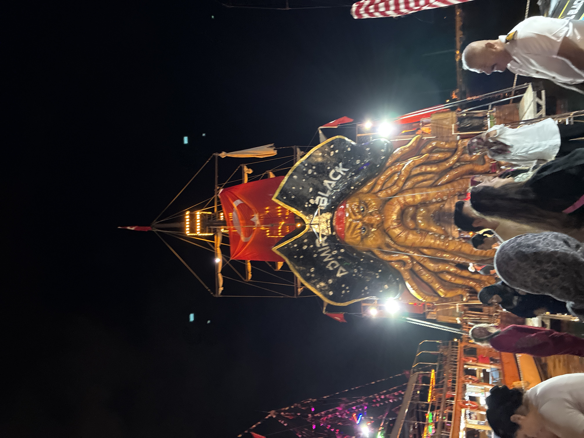 The image captures a nighttime scene at what appears to be a fairground or entertainment venue in Alanya, Turkey. The primary subject is a large, elaborate pirate-themed attraction, likely a ride or a showpiece. It features a golden, tentacled creature resembling a kraken or sea monster, with a pirate hat and a Jolly Roger flag. The words "ADMIRAL BLACK" are visible on a black banner with white stars, part of the pirate theme. The attraction is illuminated with bright lights, including strings of yellow bulbs and spotlights, creating a festive and somewhat dramatic atmosphere.
In the foreground, several people are visible, mostly blurred due to the low light and movement. Some are looking towards the attraction, while others are engaged in conversation. The people are dressed in casual attire, suggesting a relaxed evening outing. One man in a white shirt is prominently featured in the upper right, appearing to look down at the attraction. Another individual in a red jacket is seen in the lower right.
The background is dark, indicating it is nighttime. The presence of other structures, possibly part of a boardwalk or amusement park, can be seen with more lights and decorative elements, including strings of colorful pennants and lights. The overall mood conveyed is one of excitement and entertainment, typical of a lively evening at a tourist destination. There are no clear weather cues, but the nighttime setting suggests it is not actively raining. The location is identified as Alanya, Turkey, which aligns with the festive, tourist-oriented vibe.