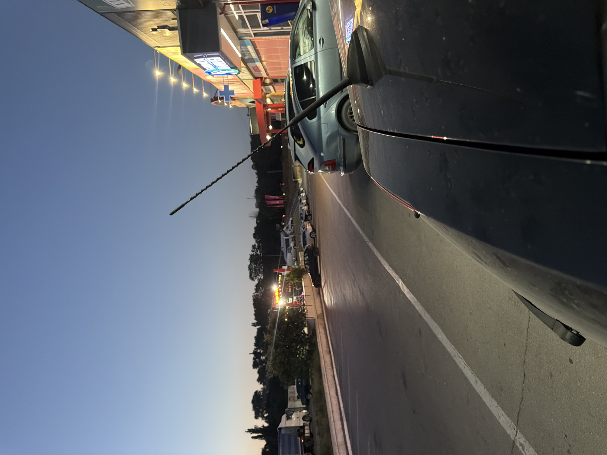 The image captures a street scene at dusk in Orhei, Moldova, with a focus on a row of parked cars and commercial buildings. In the foreground, the hood and windshield of a dark-colored car are visible, angled towards the right. To the left, a light blue car is parked on a paved surface, with a tall, thin antenna extending upwards from its roof. Behind this car, other vehicles are parked in a line.
The background features a modern building with illuminated signage, including bright lights along its roofline and a neon blue cross symbol. To the far left, a large white truck is parked, accompanied by other vehicles and some trees. The sky is a gradient of pale blue, transitioning to a darker hue at the top, indicating twilight. The overall setting appears to be a commercial area or parking lot on the outskirts of the city. No people are visible in the frame.