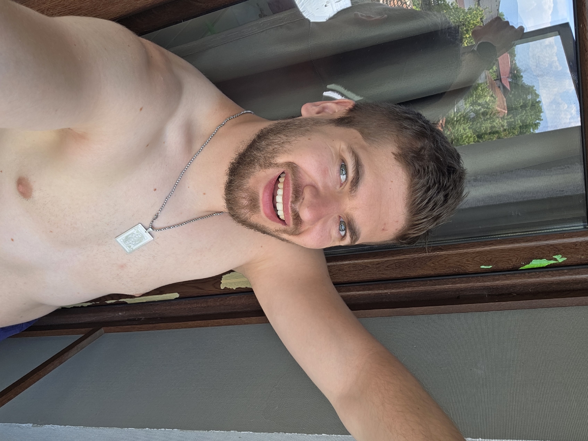 The image captures a shirtless male individual taking a selfie in Yagodovo, Bulgaria. The male has fair skin, short light brown hair, blue eyes, and a brown beard and mustache. He is smiling broadly, revealing his teeth. He wears a silver chain necklace with a rectangular silver pendant featuring an intricate embossed design. His left arm (viewer's right) is raised and extended to the right. To the immediate right of the individual (viewer's left), a dark brown wooden window or door frame is visible, bearing chipped bright green paint residue. A reflective surface within this frame reveals a distorted reflection of the individual's back and the hand holding the camera. Through the window, an outdoor landscape is seen under a blue sky with scattered white clouds, featuring green trees, red-tiled roofs of residential structures, and distant hills or mountains. To the left of the individual (viewer's right), a light gray, horizontally textured surface, possibly a wall or mosquito netting, is visible. The scene is illuminated by natural daylight.