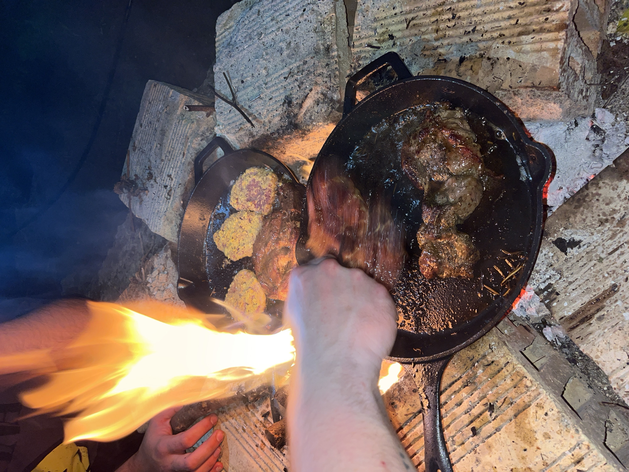 The image shows two black cast iron pans set on a makeshift outdoor cooking setup constructed from concrete blocks, possibly in Varna, Bulgaria. One pan contains what appears to be patties coated in yellow breadcrumbs, while the other holds several browned pieces of meat simmering in oil. A person's arm and hand reach into the scene, presumably to manipulate the food in the pans. Flames are visible, rising towards the left pan and between the arm/hand. The environment is dimly lit, suggesting night or a covered space. The ground surrounding the cooking setup is dirty, with ash and debris present near the edges of the concrete blocks.