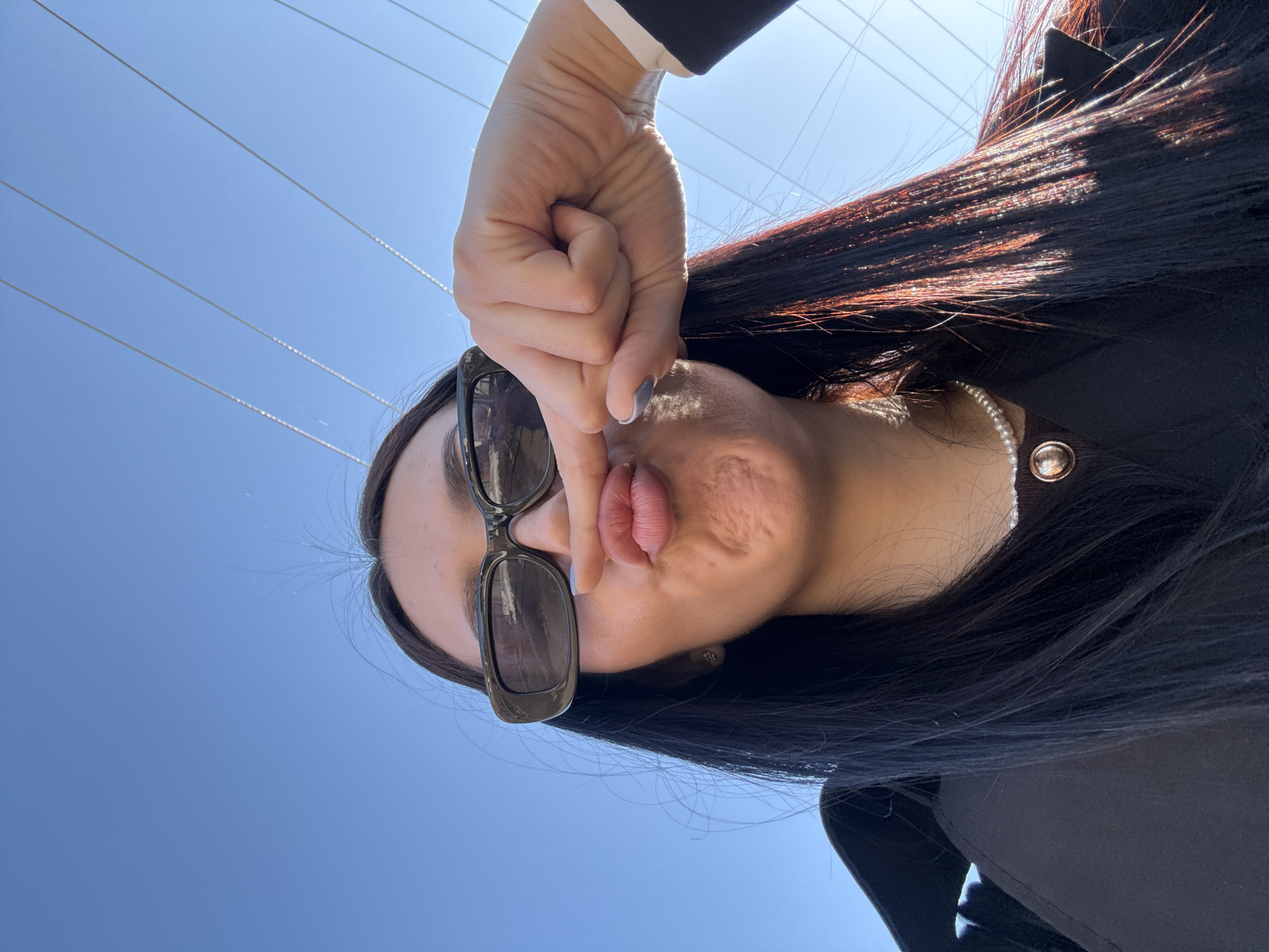 A woman with dark hair and sunglasses is taking a selfie outdoors against a bright blue sky with visible power lines. She is making a face, pursing her lips and holding her hand near her face. She's wearing a black blazer and a pearl necklace. It is impossible to determine if the location is in the city of Votkinsk, Russia based on the information provided.