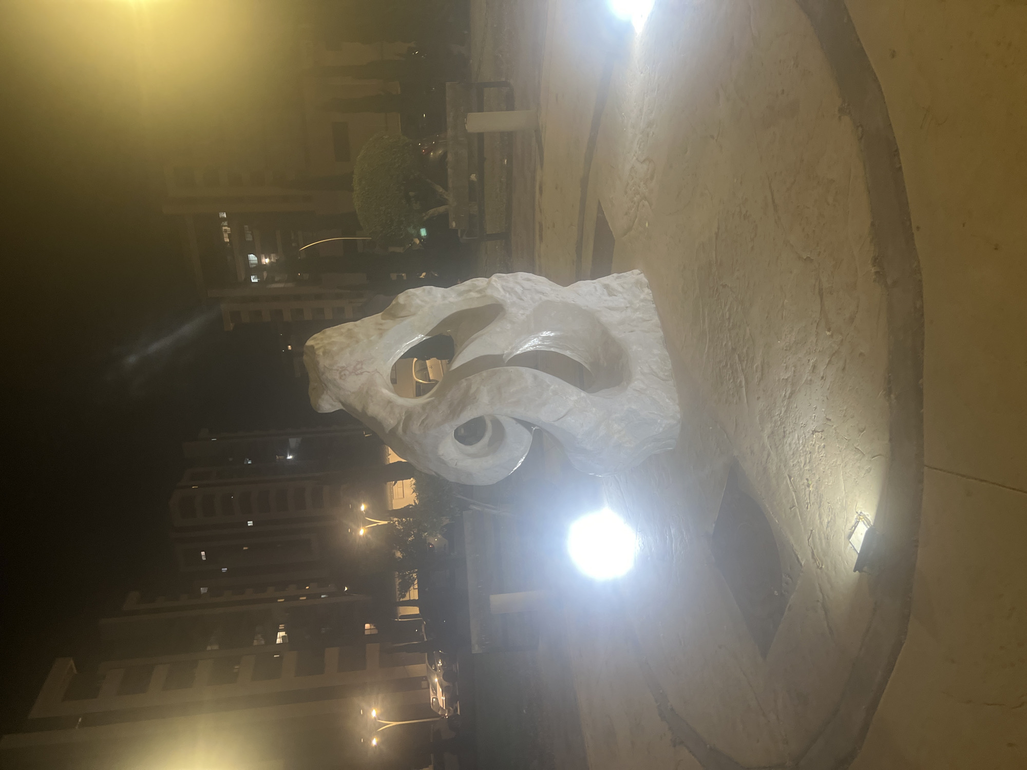The image shows a white, abstract sculpture in a public park at night in the city of Or Akiva, Israel. The sculpture is roughly human-shaped but with a flowing, organic design, featuring several openings and curved surfaces. It is lit by spotlights from below, creating a dramatic effect and casting shadows. In the background, apartment buildings are visible, illuminated by interior lights. Streetlights and parked cars are also present, suggesting the park is located near a residential area. The ground around the sculpture is paved with light-colored stone, and there is a circular path surrounding it. A bench and some trees are visible in the middle ground. The overall atmosphere is calm and serene, with the lighting highlighting the artistic elements of the sculpture and its surroundings.