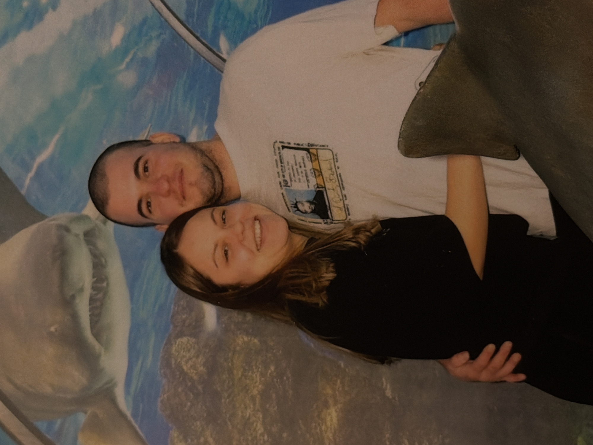 A photograph depicts a man and a woman in an embrace, likely at an aquarium. The man has a shaved head, light skin, and a short beard, and he is wearing a white t-shirt. The woman has long brown hair and is wearing a black top. Behind them is a backdrop showing an underwater scene, including a dolphin and a shark. The picture appears to have been taken in an artificial environment, likely a tourist attraction in Varna, Bulgaria.