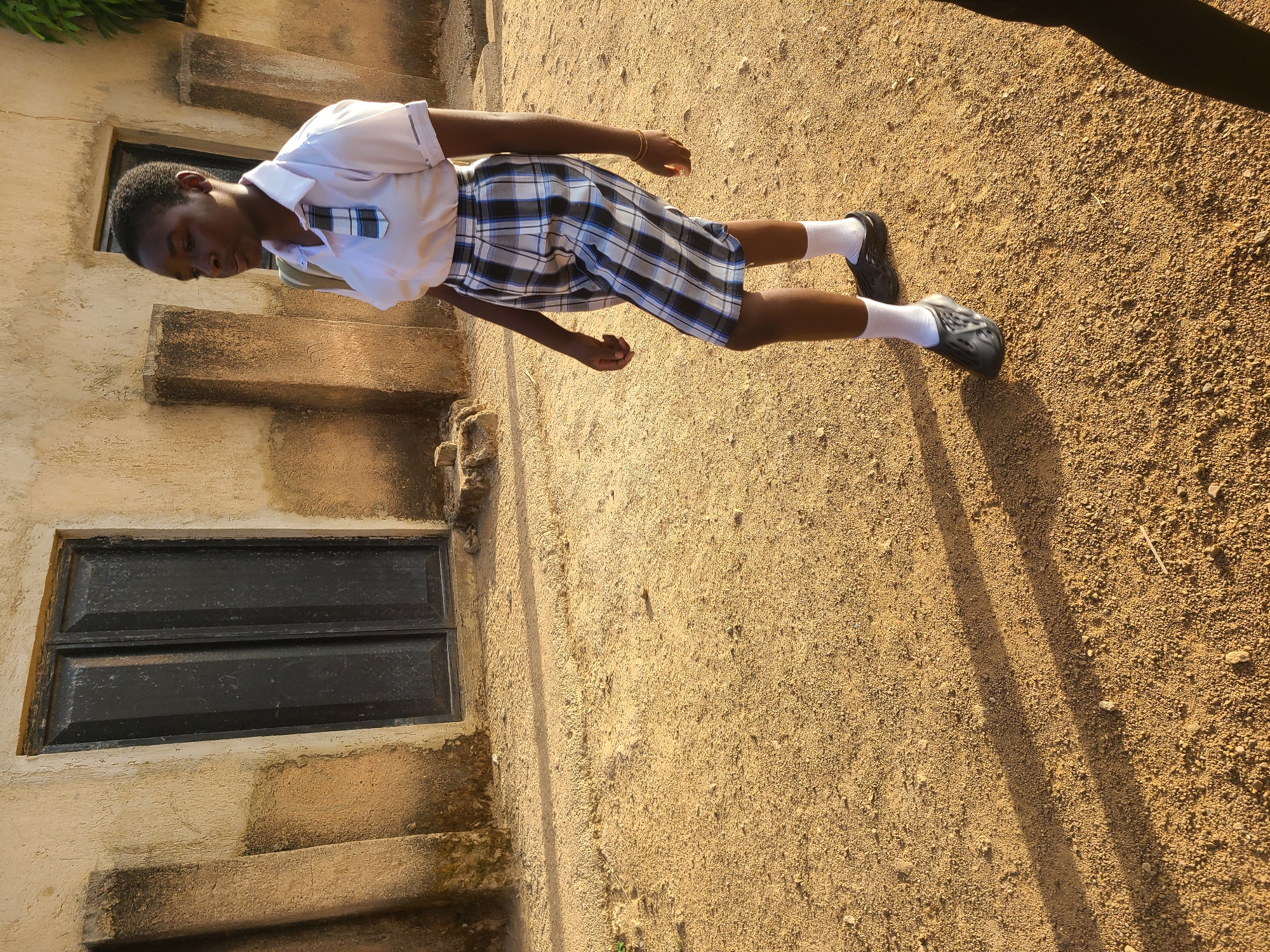 A young person stands on a dirt surface next to a building with a window and steps. The person is wearing a white shirt, a plaid skirt, white socks, and black shoes. The setting appears to be Barakin Lerawi, Nigeria.