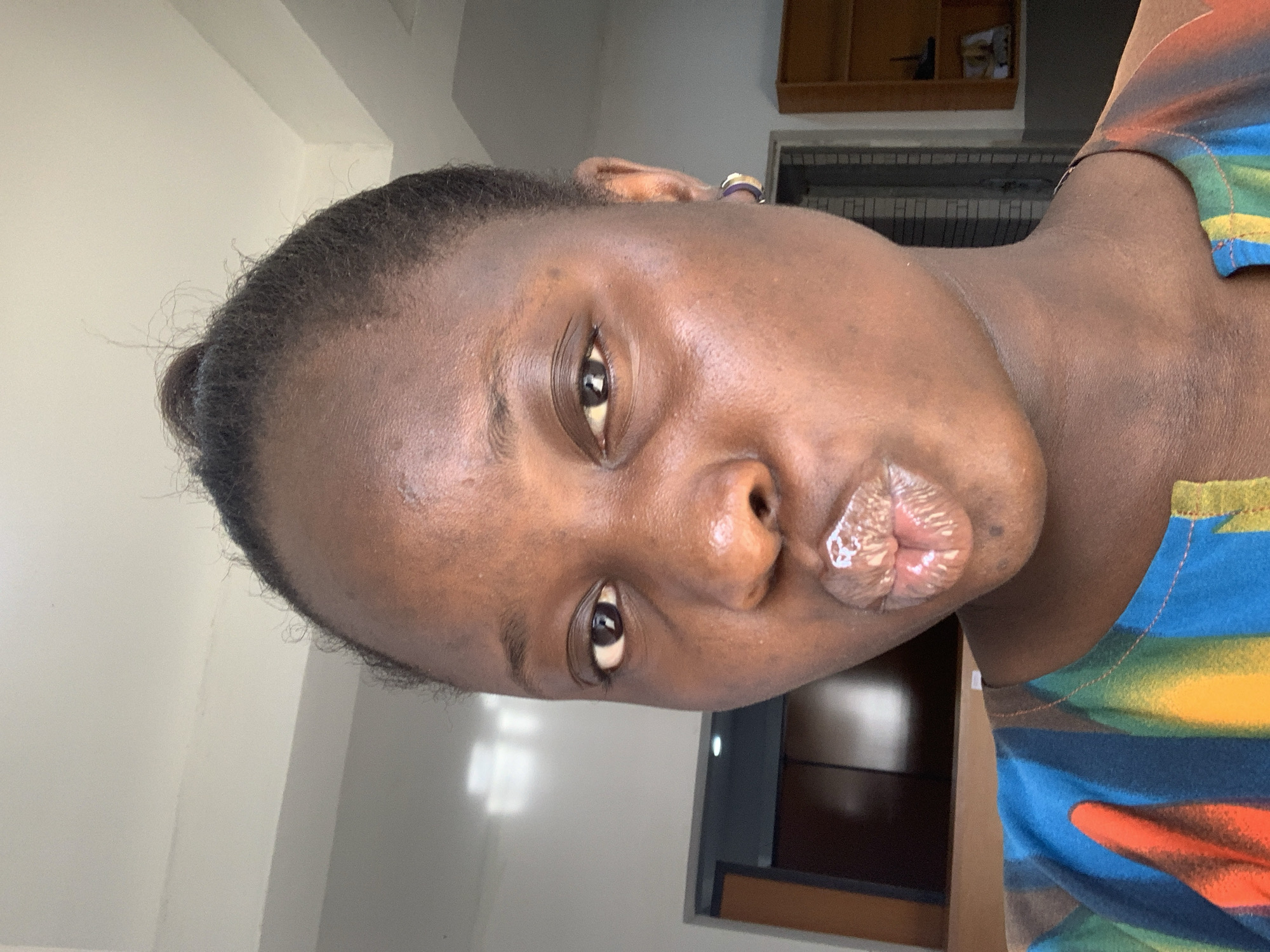 A close-up selfie shows a young woman with dark skin, her face angled towards the camera. She has her hair pulled up in a bun, and is wearing a colorful, striped shirt. Her lips are pursed and shiny, with gloss or balm visible. A small, dark earring is visible on her right ear. The background suggests an indoor setting, possibly a room with light-colored walls and ceiling, and a hint of furniture or shelving in the distance. The lighting is bright, casting subtle shadows.
