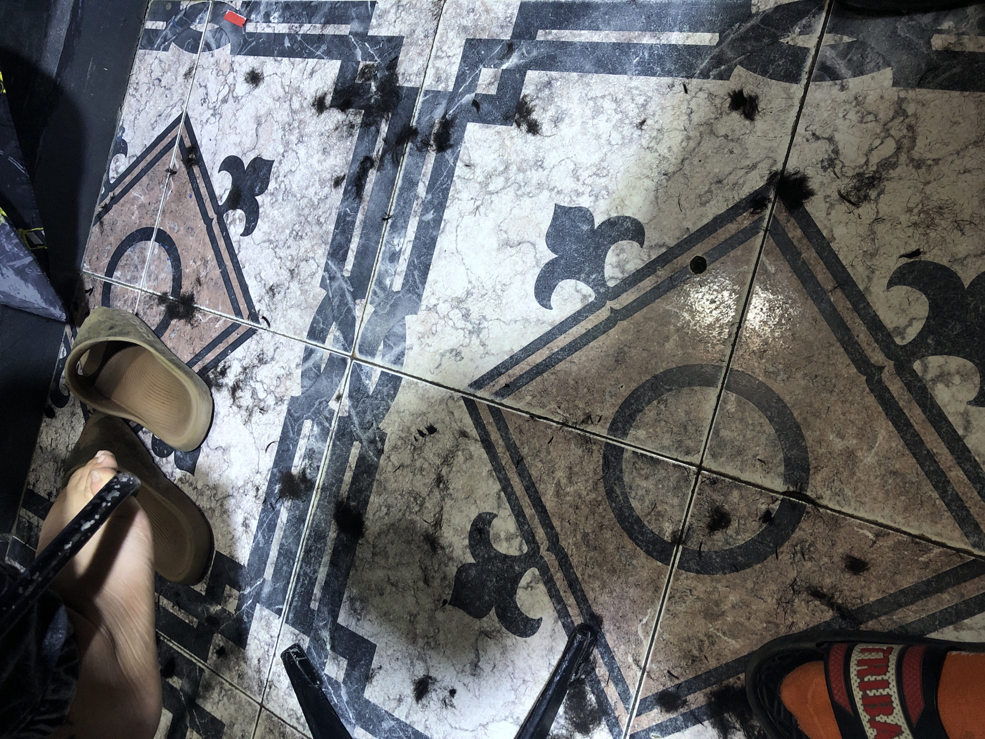 The image depicts a section of tiled floor. The tiles are decorated with a repeating geometric pattern including circular and fleur-de-lis motifs, in a palette of tan, brown, black and white. Scattered across the surface of the tiles are dark clippings, likely hair. There are two pairs of sandals, one beige, with a foot partially visible, and the other orange and red. The image was taken indoors. There is no visual evidence in the image to confirm its location in Altagracia, Venezuela.