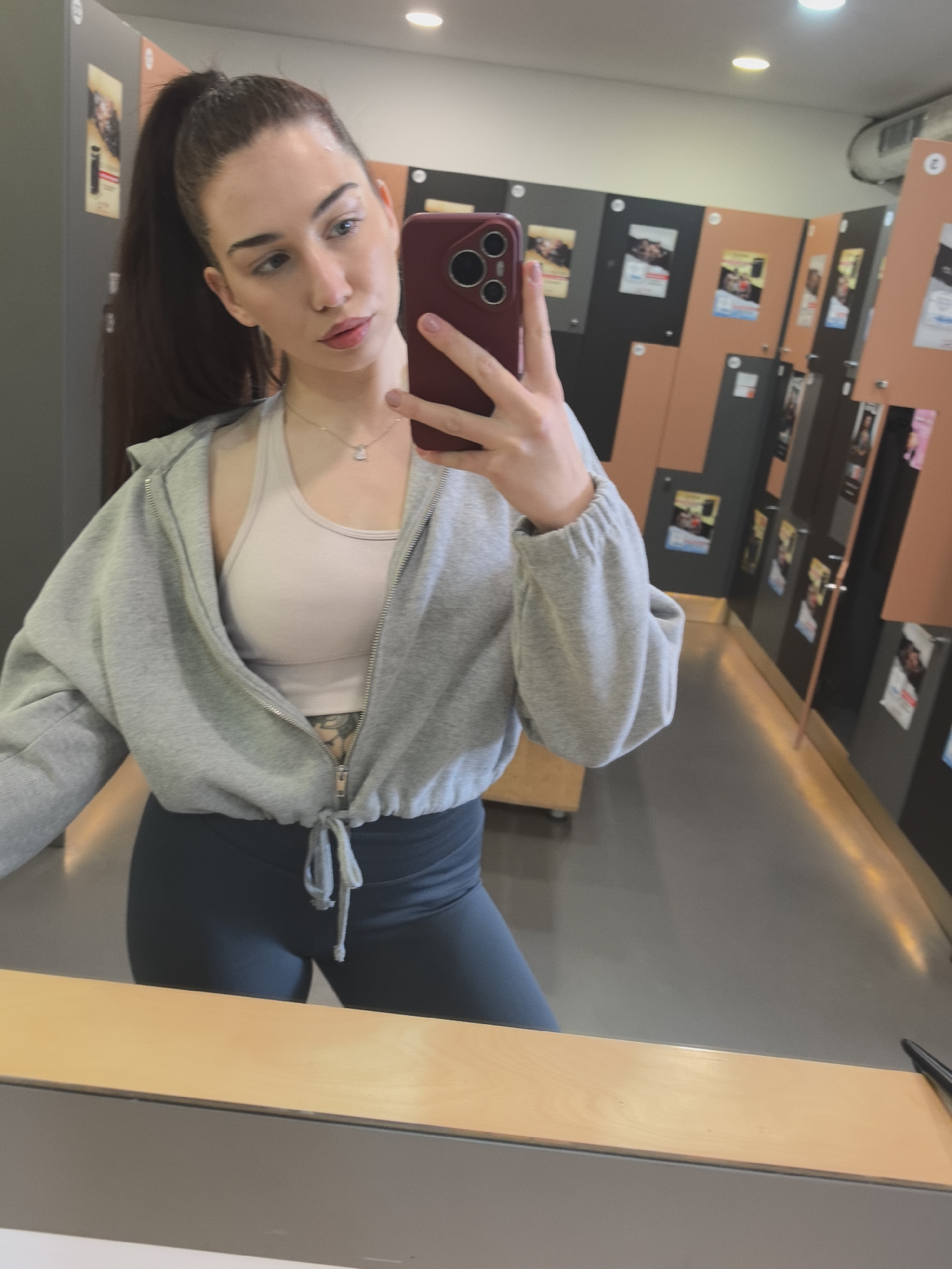 A female individual is depicted in a mirror selfie, taken within an indoor locker room or changing facility, contextually placed in Stara Zagora, Bulgaria. The subject, with dark hair styled in a high ponytail and light-colored eyes, holds a dark red smartphone in her right hand to capture the image. She wears a light grey cropped zip-up hoodie, unzipped, over a light beige sports bra, and dark-colored leggings. A silver-colored necklace with a small pendant is visible, along with a partially obscured tattoo on her left upper abdomen. The background shows a corridor lined with a series of vertically oriented lockers, featuring alternating dark grey and light brown/orange panels. Various posters or notices are affixed to the locker doors. The floor is a uniform light grey, and the white ceiling contains recessed circular light fixtures. A light wood-colored bench or counter spans the lower foreground.