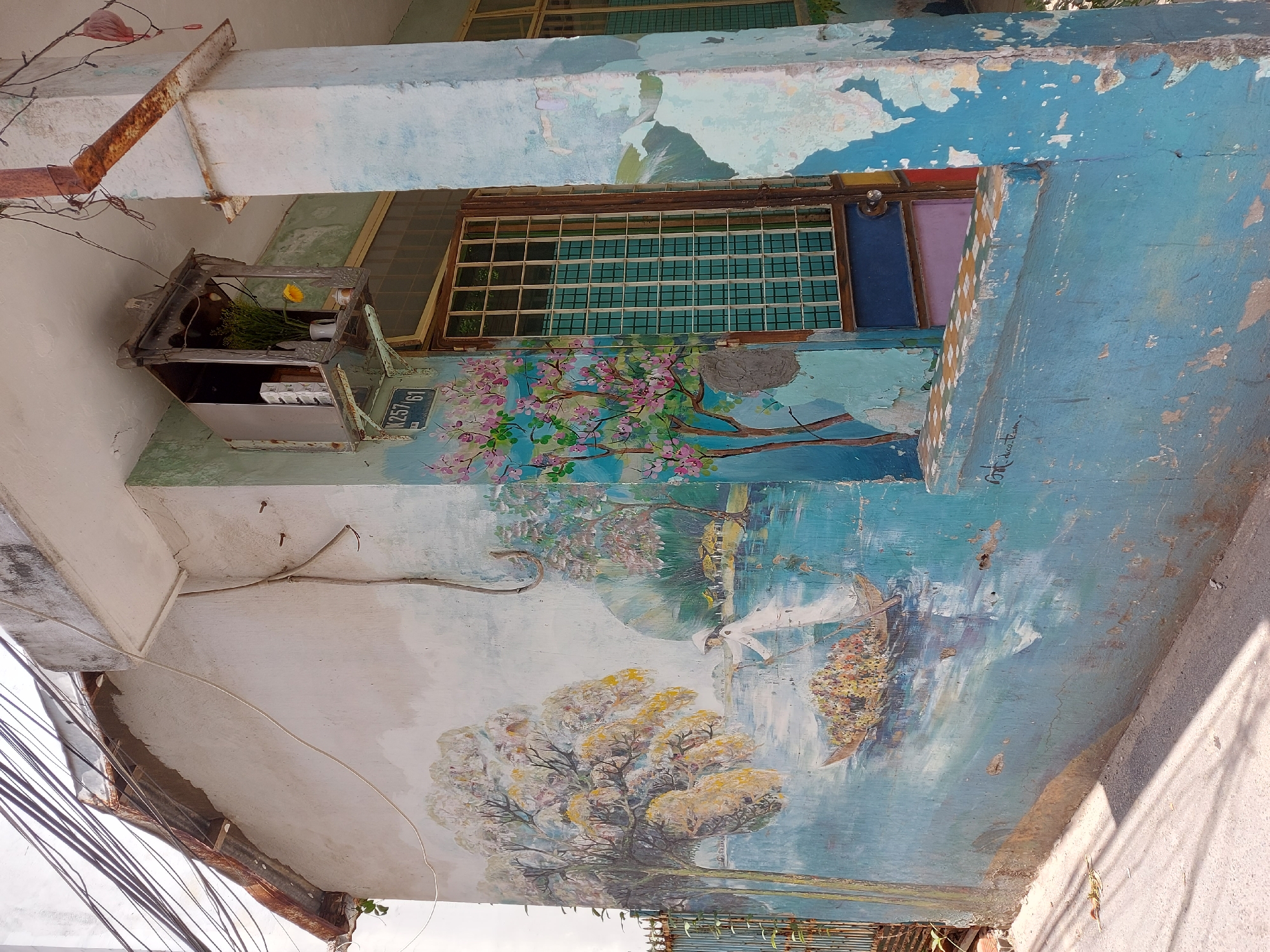 This is a photograph of a weathered building exterior adorned with a mural depicting a serene river scene in Vietnam, possibly in the city of Đà Nẵng. The mural shows a woman in traditional Vietnamese attire, wearing a conical hat and a white garment, poling a boat laden with colorful flowers through calm, blue water. In the background, lush trees with white and pink blossoms frame a distant village with thatched-roof houses. The mural extends across the wall, wrapping around a window and a concrete pillar. The building itself shows signs of age, with peeling paint and rust on a metal downspout and window bars. A small, ornate shrine, possibly for offerings, hangs above the window. Electrical wires are strung across the top of the building, indicating an urban or semi-urban setting. The ground in front of the building is a paved surface, likely a street or sidewalk, with sunlight casting shadows.