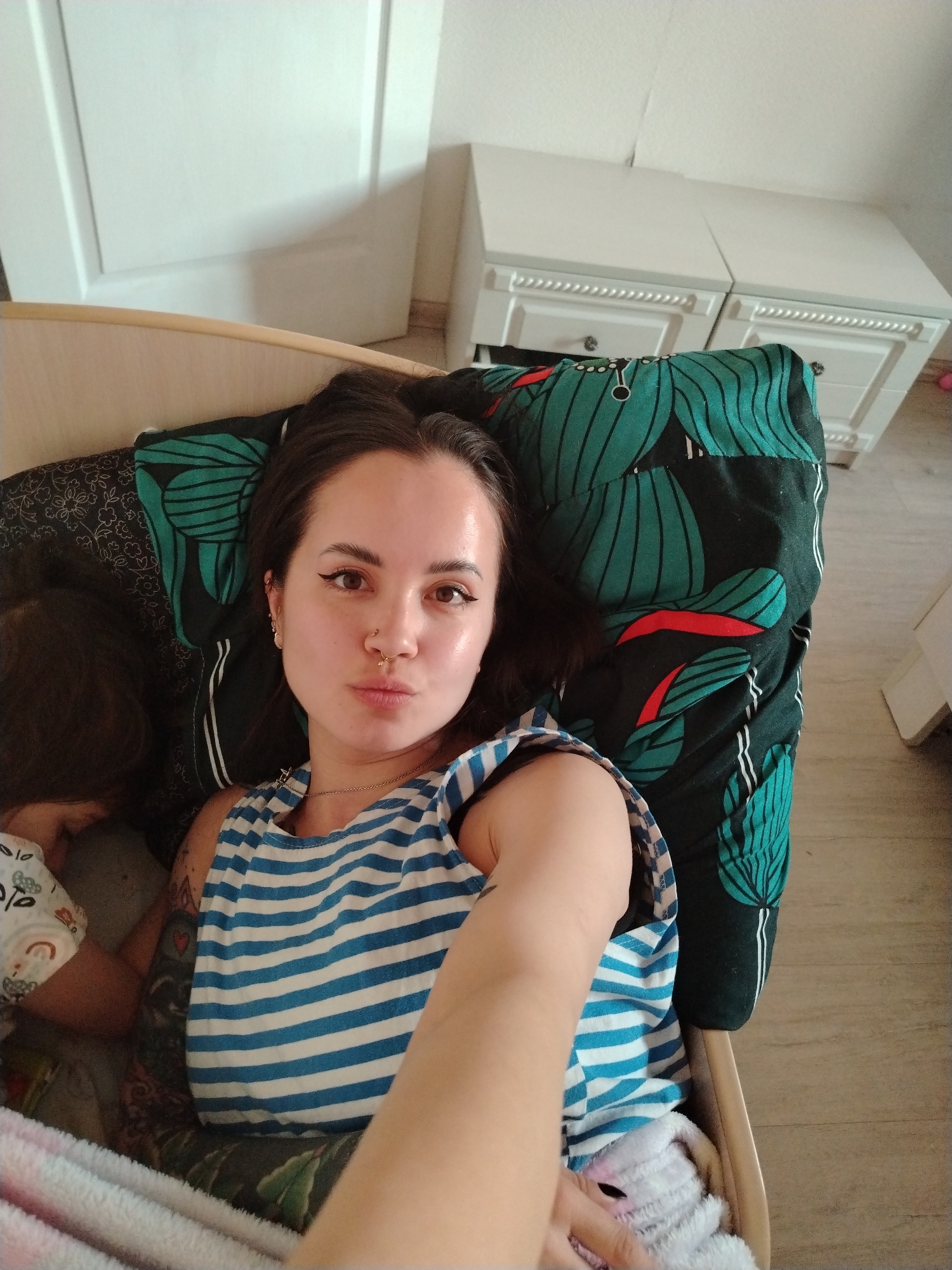 A woman with dark hair and fair skin is lying in a bed with a black and green leaf-patterned pillow. She is wearing a blue and white striped tank top and a gold necklace, and has multiple piercings in her ears and nose. A young child is lying next to her, partially obscured by the woman and bedding. The room contains a white door and two white bedside tables with drawers. The location is likely a private residence, but there is insufficient information to confirm it is located in Noviy, Russia.
