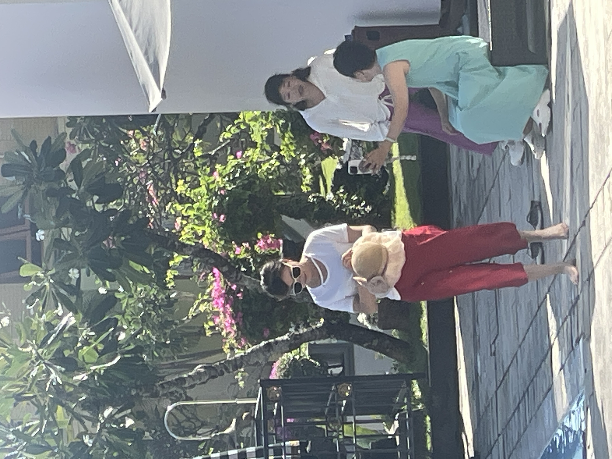 In Kuta Selatan, Indonesia, three women are interacting outdoors near a swimming pool. The woman in the foreground, wearing a white t-shirt and red capri pants, holds a straw hat. To her right, another woman in a white blouse and purple pants is talking and holding up a smartphone. A third woman, in a light blue dress, is seated on a dark-colored step next to the woman with the smartphone, looking towards her. Lush green trees with pink flowers are visible in the background, along with a building and part of a black metal fence. The ground is paved with square tiles, and the edge of a blue swimming pool is visible in the lower left corner.