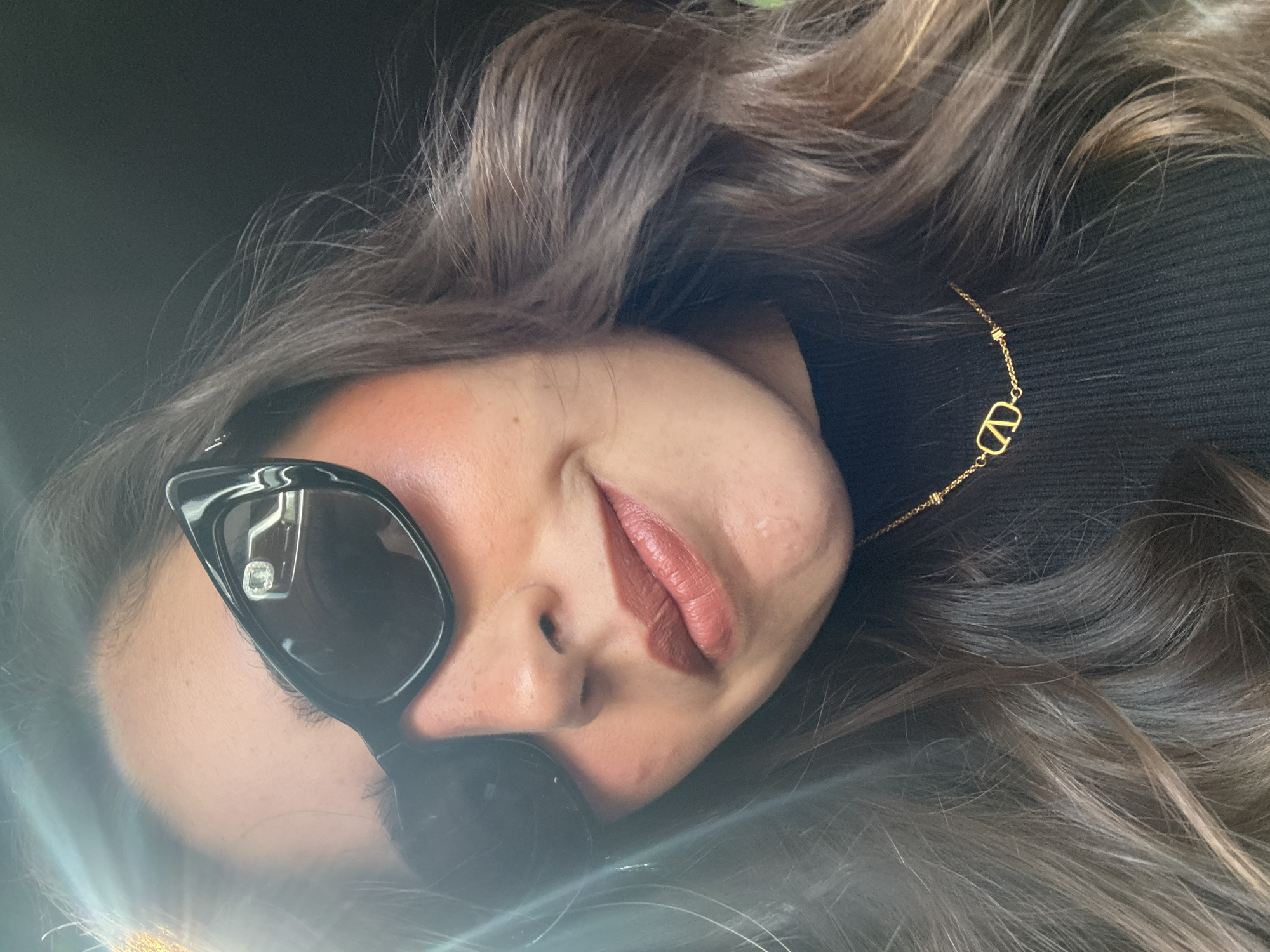 A female is captured in a close-up, slightly rotated shot. She wears sunglasses and a black turtleneck, adorned with a gold necklace featuring a "V" pendant. The background is dark and indistinct. The location cannot be determined from the image alone.