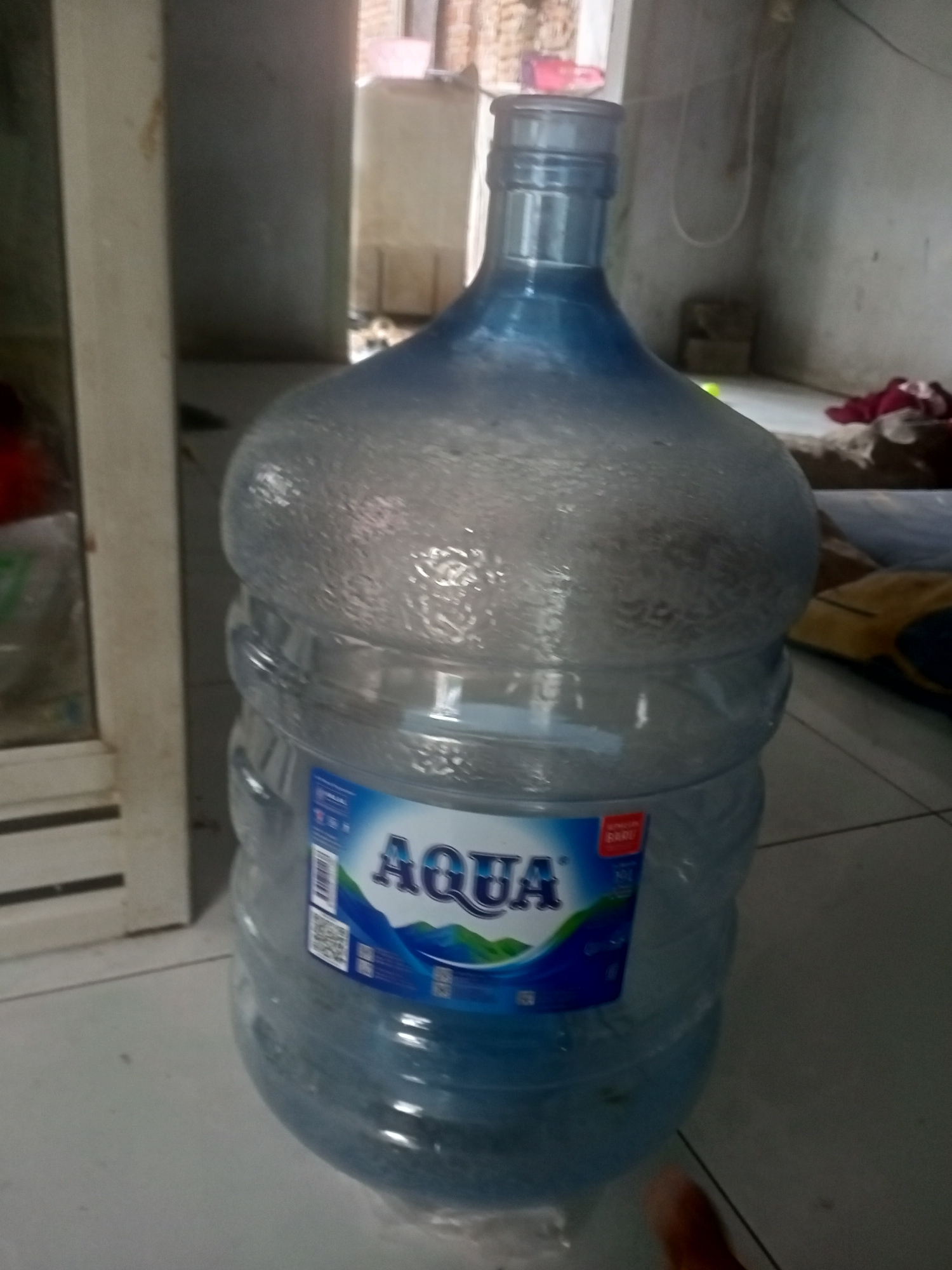 An indoor, medium shot captures a large, clear plastic water bottle, half-filled with water and labeled "Aqua," placed on a tiled floor. The bottle's blue-tinted cap and upper section are visible, as is its ridged body. The background reveals a somewhat cluttered domestic environment with a partially open door, furniture, and red and white fabric visible on the floor. The lighting appears to be natural, coming from a window or an open doorway, casting shadows and creating a slightly dim atmosphere. The image provides no specific geographical indicators for Badas, Indonesia.