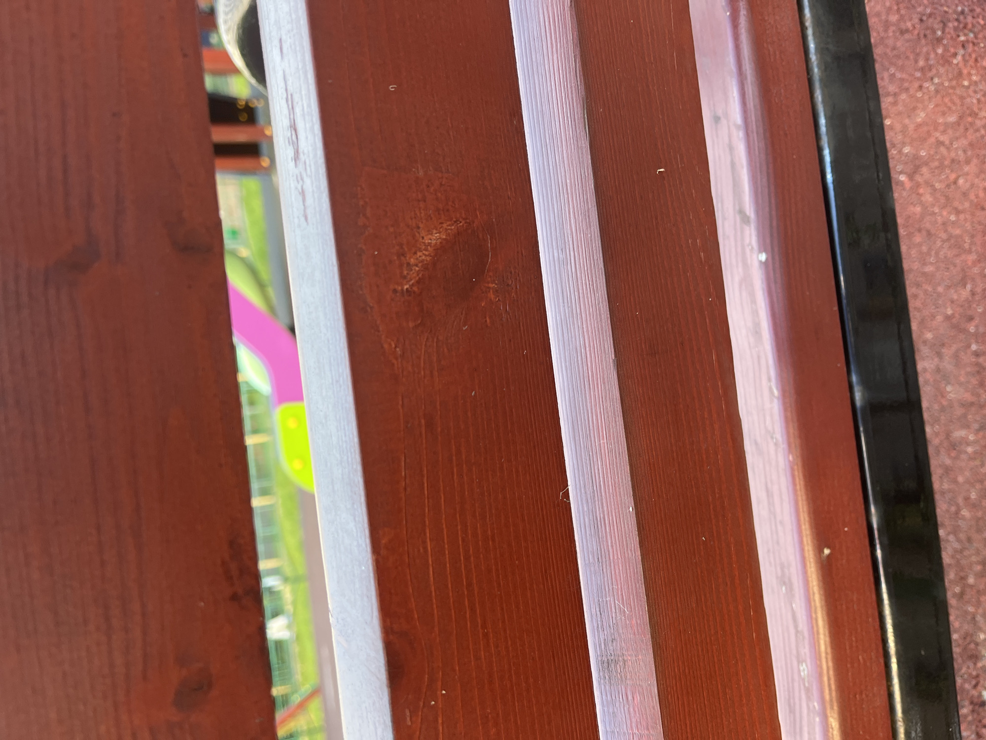 The image shows a close-up view of a dark red wooden bench. The bench has visible wood grain and appears to be outdoors, possibly in a park or playground. Through the slats of the bench, a blurred background can be seen which includes green grass, a bright pink and yellow slide, and some wooden structures. The ground beneath the bench is a reddish-brown, textured surface. The lighting suggests it is daytime. The specific location in Nemchinovka, Russia cannot be determined from the visual information alone.