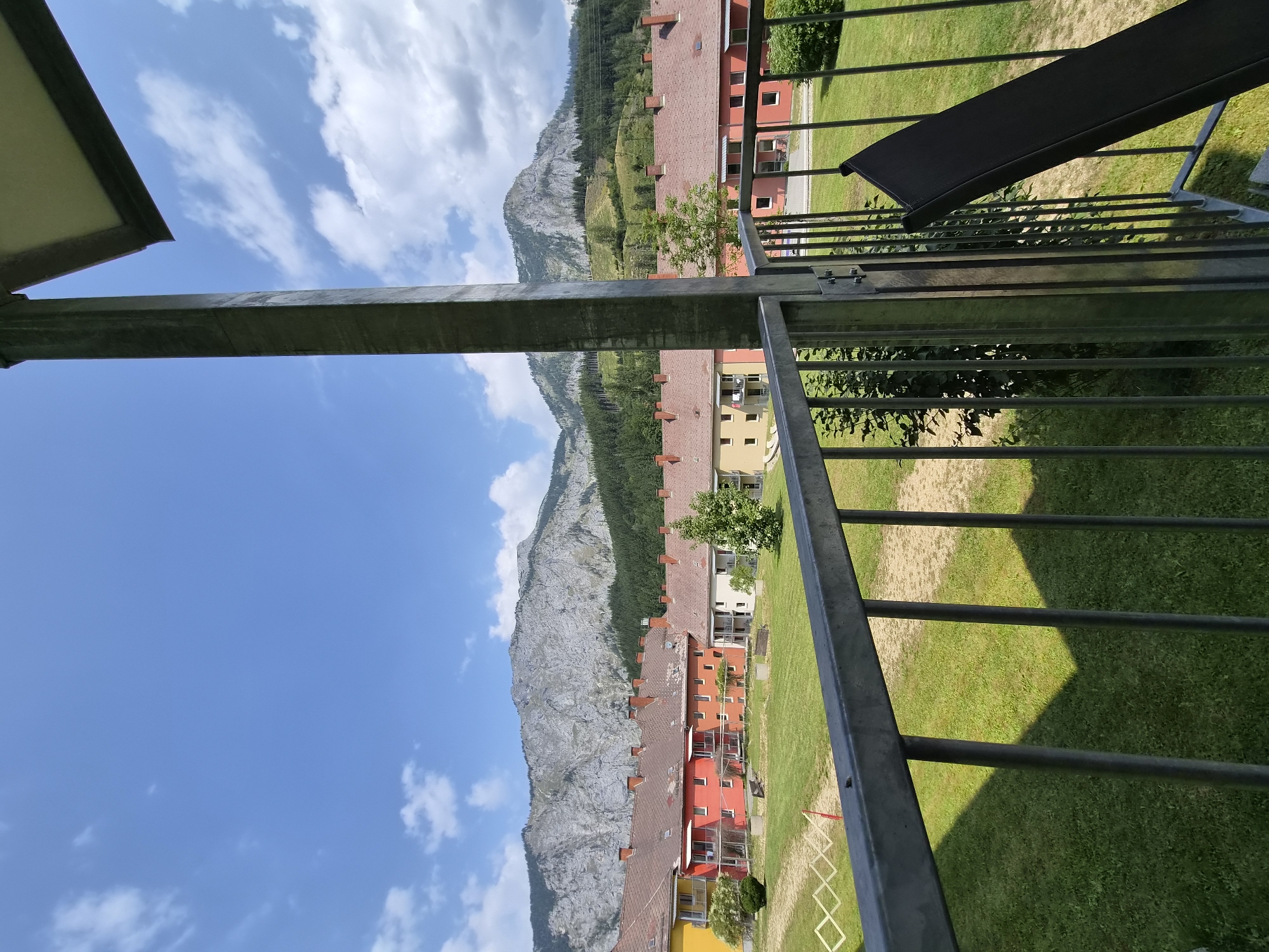The image captures a view from a balcony overlooking a residential area in Eisenerz, Austria. In the foreground, a metal railing separates the balcony from a well-maintained green lawn. A dark folding chair is partially visible, leaning against the railing. Beyond the lawn are several multi-story buildings with reddish-brown tiled roofs and brightly colored facades, predominantly orange and yellow. Behind the buildings, a lush green mountain range dominates the background, with rocky peaks and dense coniferous forests. The sky is a vibrant blue with scattered white clouds. The overall scene suggests a peaceful, sunny day in a picturesque Austrian town.
