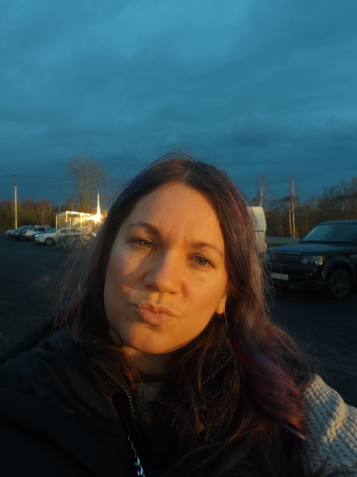 A woman is captured in a selfie outdoors, featuring her face prominently in the foreground, illuminated by warm, golden light. She has long, dark hair with subtle reddish-purple highlights and is making a playful puckering expression with her lips. She wears a dark jacket over a light-colored, possibly knitted, sweater.
The scene appears to be set during golden hour, either at sunrise or sunset, given the dramatic contrast between the warm light on the woman's face and the deep, rich teal-blue sky filled with dark clouds. In the background, there's an expansive parking area or open space. Several cars are parked on the left, and a bright light source, possibly a streetlight, casts a glow behind them. On the right, a dark SUV, identifiable as a Range Rover, is parked. Its license plate is partially visible, showing "TAT" followed by numbers. Bare trees line the horizon in the distance. The location context provided is Svistukha, Russia.