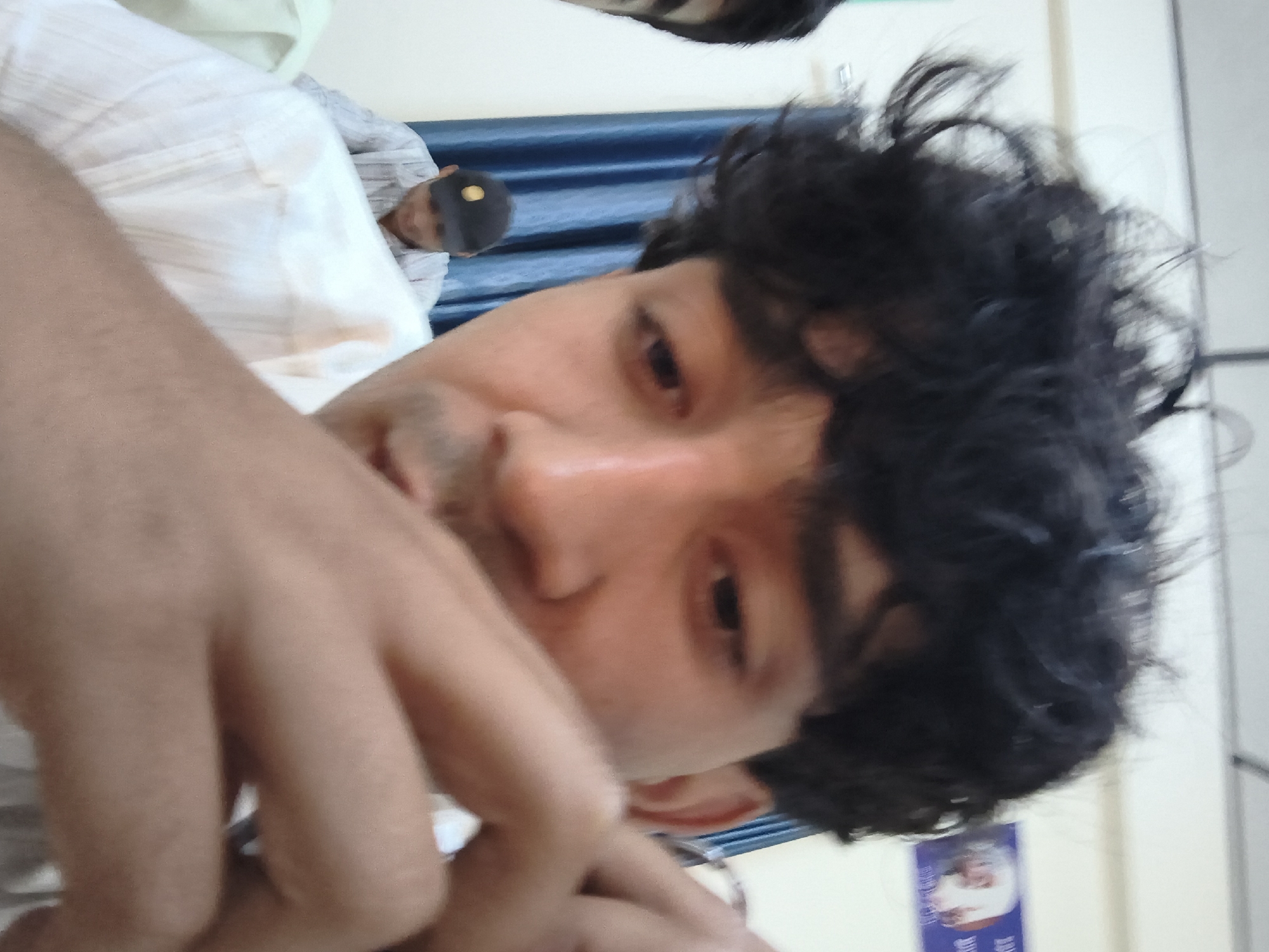 The image is a close-up, slightly blurry selfie taken indoors. The primary subject is a young man with dark, curly hair, his face partially obscured by his hand. He is wearing a light-colored collared shirt. In the background, another person wearing a dark baseball cap and a striped shirt is visible, seated. The environment appears to be a room with a white wall and a blue curtain. On the far right background, a poster with an image of a person is affixed to the wall. There is no discernible information within the image to suggest a specific location within Dharamshala, India.
