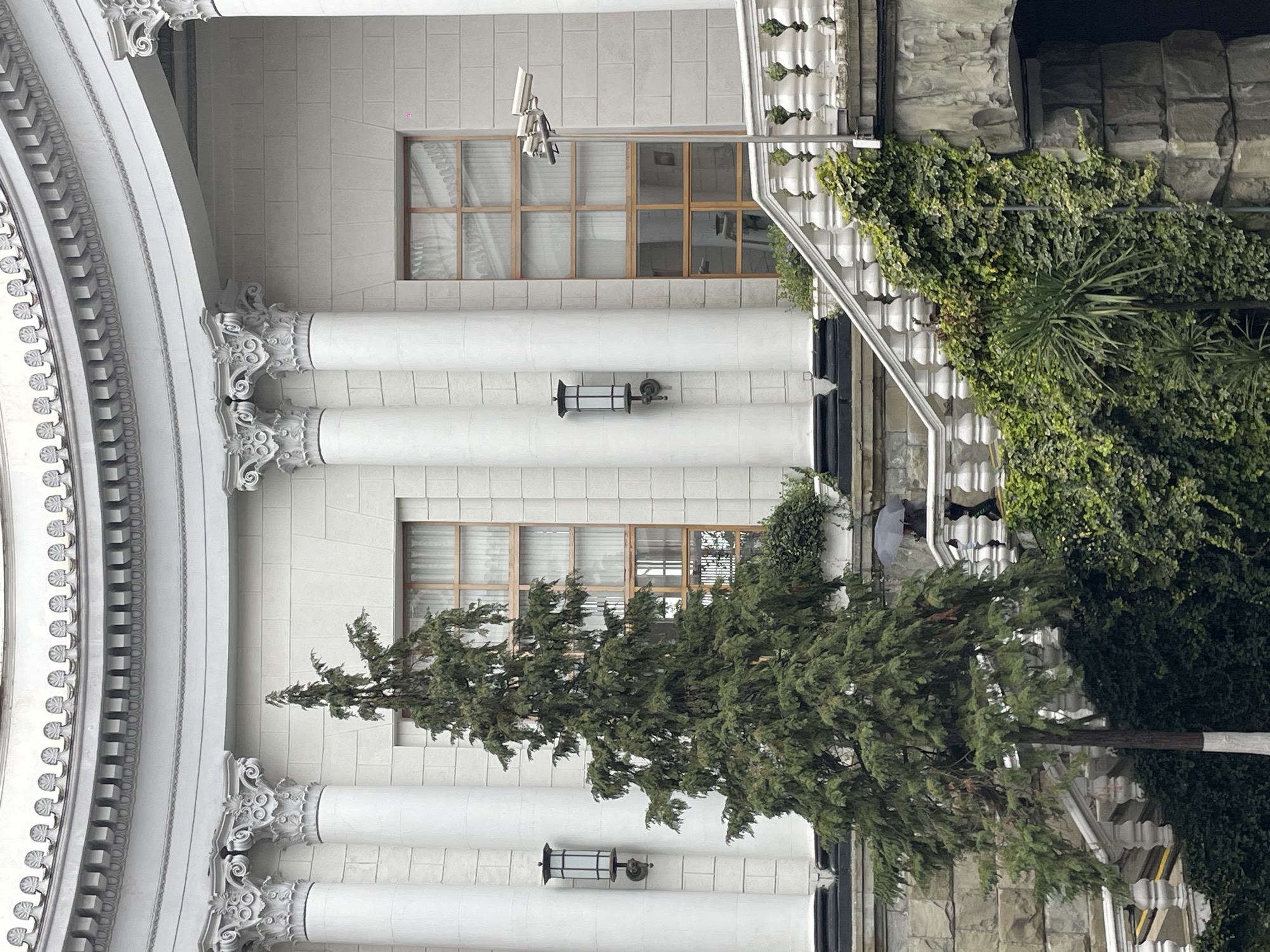 The image depicts a section of a building facade, likely in Sochi, Russia, based on architectural styles. The building features classical elements such as ornate columns, a decorative arch, and rectangular windows. The exterior walls are composed of light-colored stone or concrete blocks.
A surveillance camera is mounted above one of the windows. Outdoor lighting fixtures are attached to the wall between columns.
To the right of the building facade, there is a stone wall covered in dense greenery, including ivy and some palm-like plants.
A tree is planted in front of the building. The tree's trunk is partially visible.
A person holding an umbrella is visible between the plants on the right side of the image. The photograph appears to have been taken from a ground-level perspective.