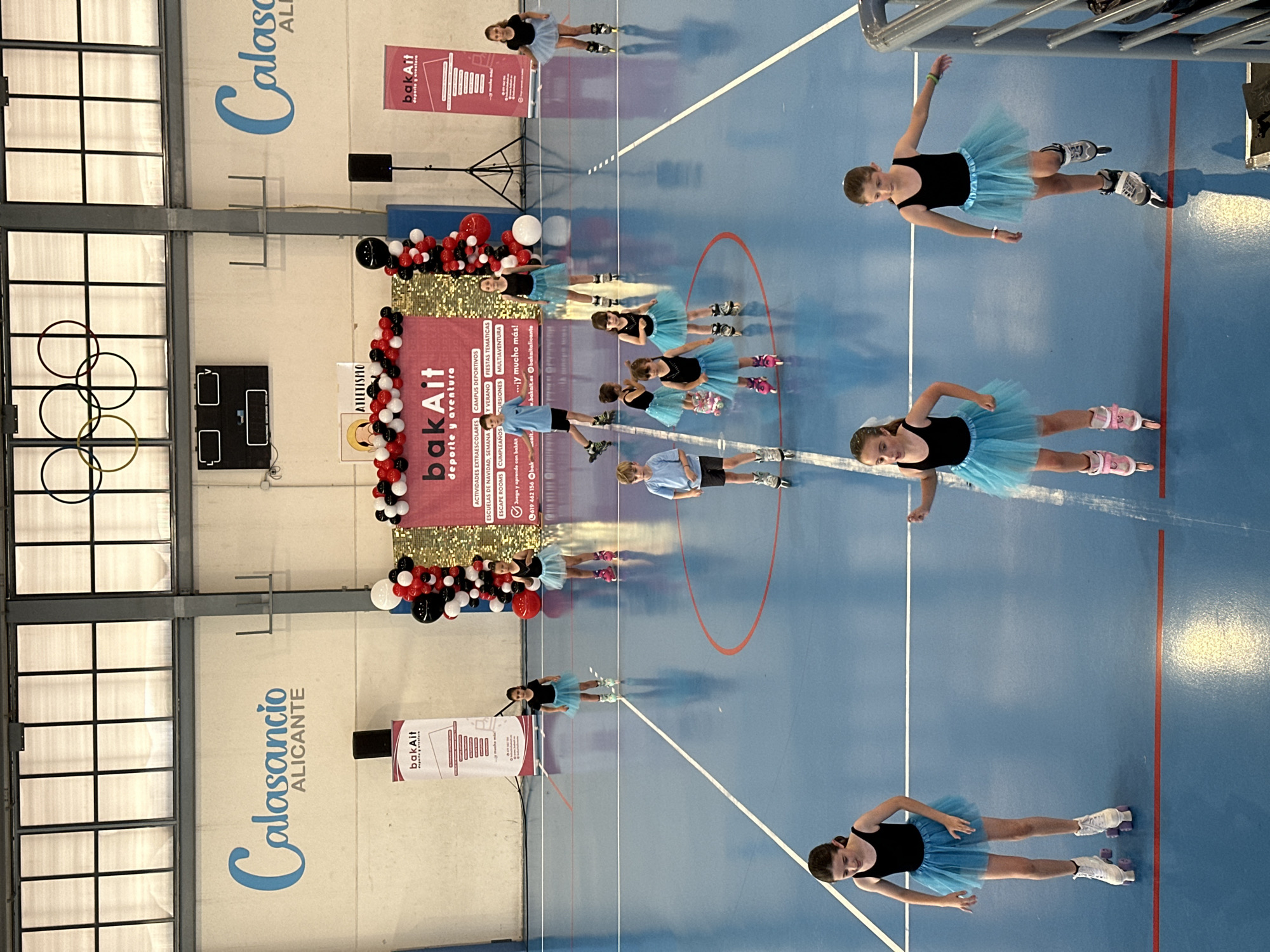 The image captures an indoor sports hall, likely a gymnasium, with a blue floor marked with white and red lines. The background wall is light-colored, featuring large window-like structures and prominent blue text that reads "Calasancio ALICANTE".
The scene depicts several young children, mostly girls, participating in what appears to be a roller skating performance or event. Most girls wear black leotards or tops paired with light blue tutus and various types of roller skates, including inline and quad skates. They are positioned in different poses on the court, suggesting a choreographed routine. Some boys are also visible, dressed in athletic t-shirts and shorts, also on roller skates.
A central backdrop features the branding "bakAit deporte y aventura" in red and white, flanked by decorative balloons in red, black, and white, indicating a festive or celebratory occasion. The backdrop lists various services offered by "bakAit," including "ACTIVIDADES EXTRAESCOLARES," "ESCUELAS DE NAVIDAD, SEMANA," "ESCAPE ROOMS," "CUMPLEAÑOS," "CAMPUS DEPORTIVOS Y VERANO," "FIESTAS TEMÁTICAS," "CURSIONES," and "MULTIAVENTURA." Contact information like "619 462 156," "w.bakait.es," and "bakaitalicante" (social media) are also visible. Another sign near the top reads "ATLETISMO." The children appear focused and engaged in their activity, contributing to an energetic atmosphere.