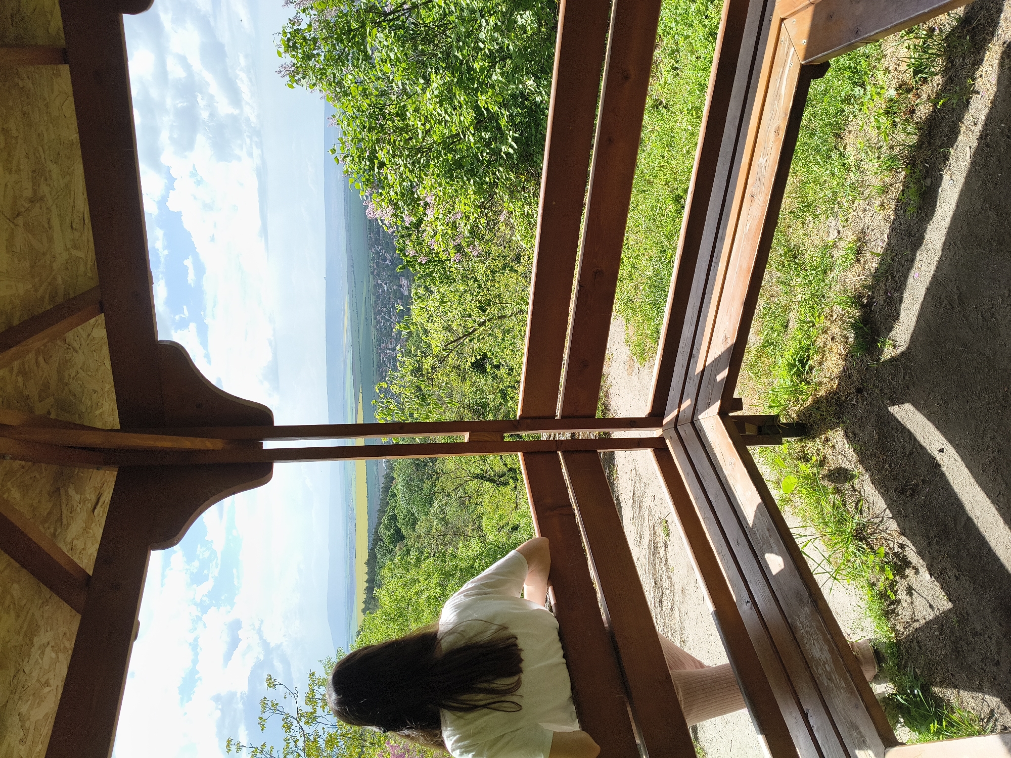 A gazebo located in Madara, Bulgaria, provides a scenic overlook. A woman with long, dark hair, wearing a white shirt and pink socks, sits on a wooden bench inside the gazebo. The gazebo has a light-colored plywood ceiling and open sides with wooden railings. The view from the gazebo encompasses green trees, grass, fields, and distant hills under a partly cloudy sky. The ground around the gazebo is covered with dirt and grass, with shadows cast by the structure.