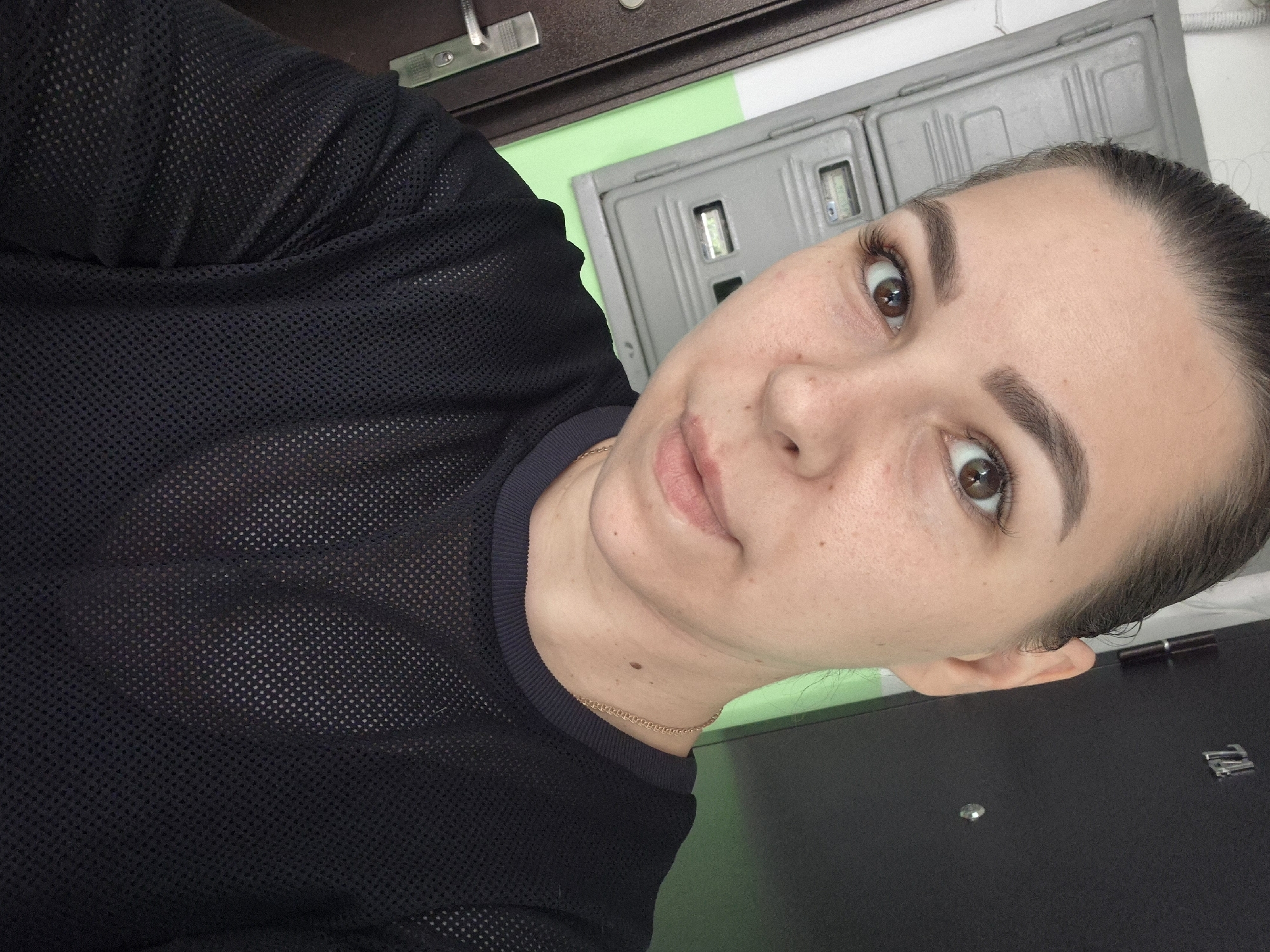 The media file is a close-up, self-portrait of a young woman. She has brown eyes and dark hair pulled back. She is wearing a black mesh top. The background is indistinct, with some elements suggesting an indoor setting with a light green wall and metallic gray panels. There are also hints of dark furniture or a door. The lighting appears to be artificial, suggesting the photo was taken indoors. The woman's expression is neutral. There is no visible text in the image. The metadata indicates the location context is Chaikovsky, Russia. The time of day and weather are not discernible from the image.