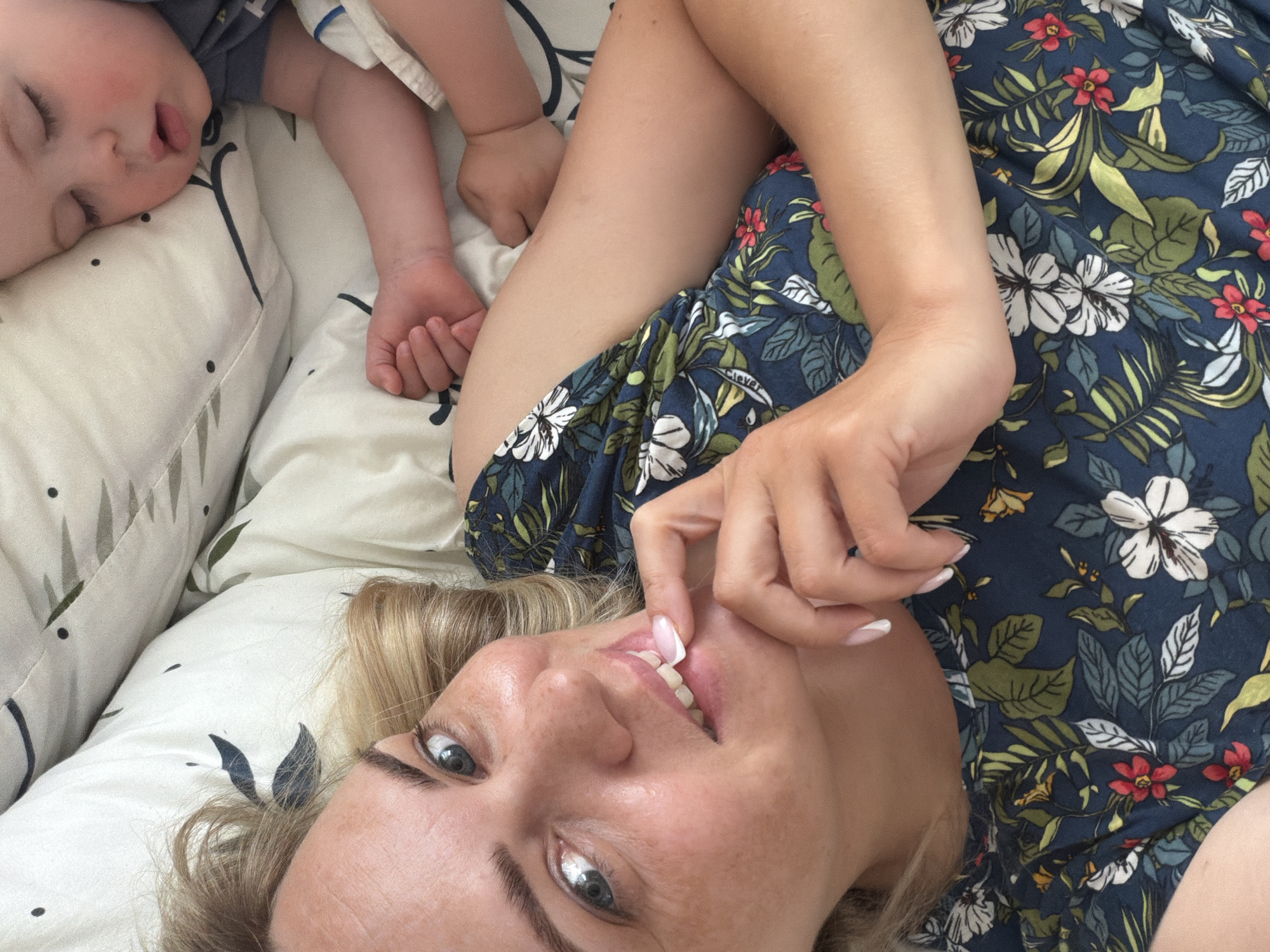 A woman with blonde hair and blue eyes lies on her back on a bed. She is wearing a dark blue floral-patterned shirt. A child with fair skin is sleeping next to her on a white pillow and blanket with a leaf pattern. The child's arm is outstretched. The woman is smiling and touching her teeth with her fingers. The location is unknown, but given the instructions, it is said to be in Votkinsk, Russia.