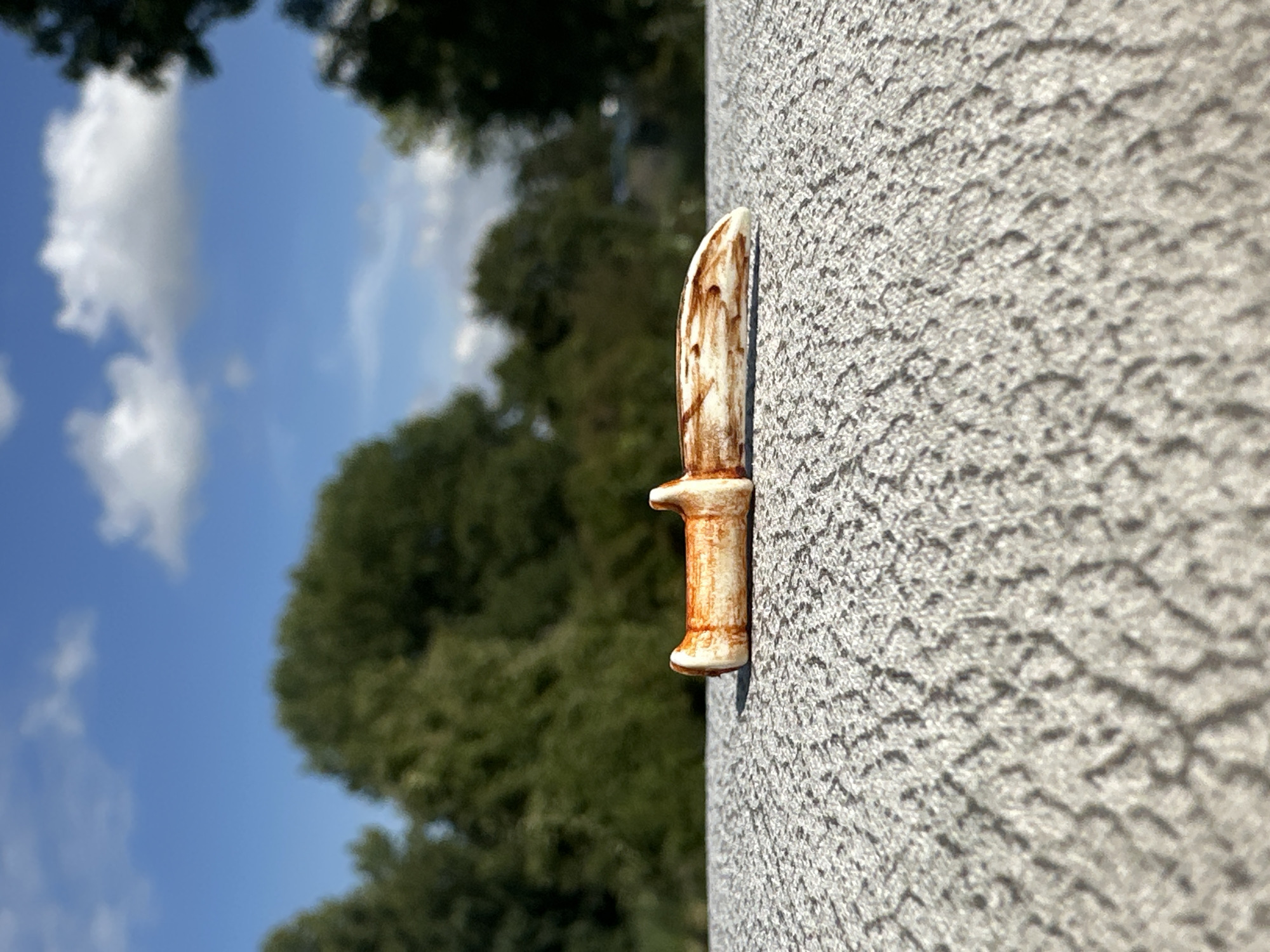 A miniature ceramic knife, approximately 5cm in length, rests on a textured, light grey surface. The knife is off-white with brown markings that resemble wood grain. The handle has a distinct hilt. The background consists of lush green trees and a bright blue sky with scattered white clouds. The scene appears to be outdoors, possibly on a patio or balcony. The image was captured in Lelystad, Netherlands.