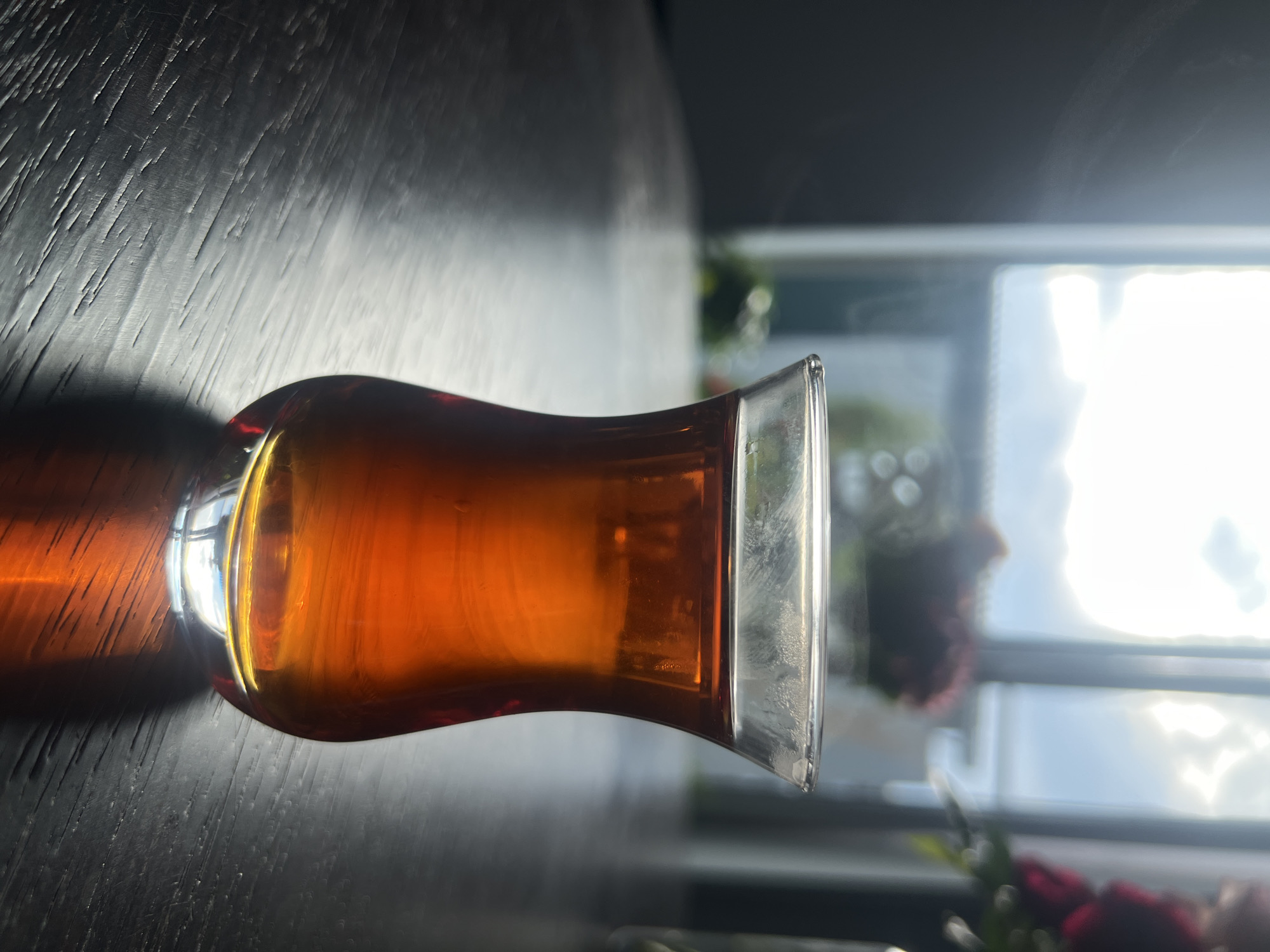 A glass of amber liquid, likely tea, sits on a dark, textured surface, possibly a wooden table. The glass has a curved shape, wider at the bottom and tapering towards the top with a flared rim. The liquid within is clear and luminous, catching the light and casting a shadow on the table. The scene is illuminated by natural light coming from a window in the background, which is bright and slightly overexposed, suggesting it's daytime. Outside the window, blurred elements of greenery and possibly flowers are visible, along with a glimpse of the sky. The overall mood is serene and contemplative, with the play of light and shadow creating a dramatic effect. There is no visible text.
