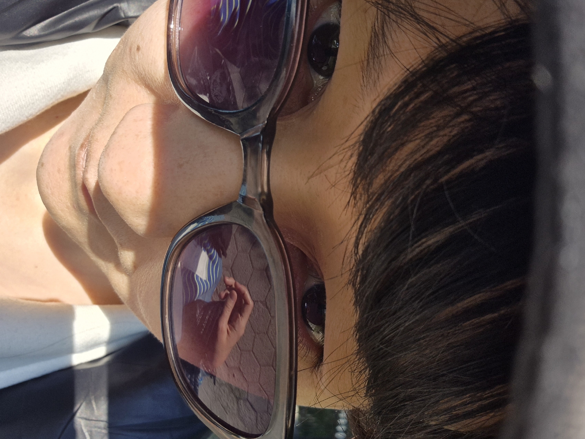 A close-up shot of a person wearing sunglasses. The reflection in the sunglasses shows a hand against a light-colored hexagonal tiled ground. The person's skin tone is fair. Hair is dark brown. The location and city are not clearly evident in the image.
