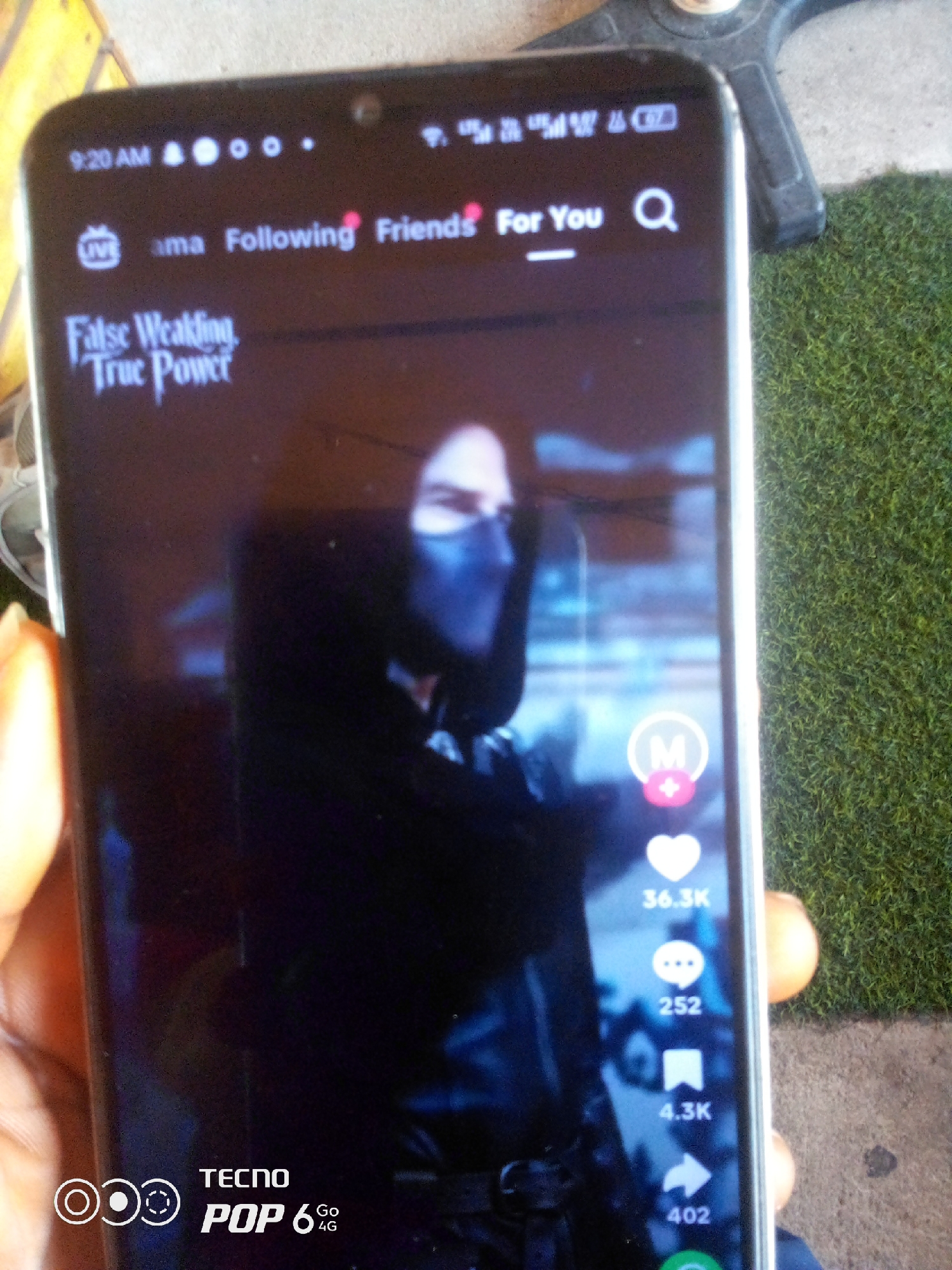 The image shows a person holding a TECNO POP 6 4G smartphone, displaying a TikTok video. The video features a person wearing a mask and dark clothing. The phone's screen shows the TikTok interface with options like "For You," "Friends," a search icon, a heart icon with "36.3K" likes, a comment icon with "252" comments, a bookmark icon with "4.3K" saves, and a share icon with "402" shares. The phone's status bar at the top displays the time as "9:20 AM" and network signal indicators. The background appears to be an outdoor setting with green turf and some yellow and grey elements, possibly a street or a paved area. The presence of the TECNO POP 6 4G branding suggests this scene could be within Nigeria. However, no specific location in Howayi, Nigeria, can be determined from the visual information provided.