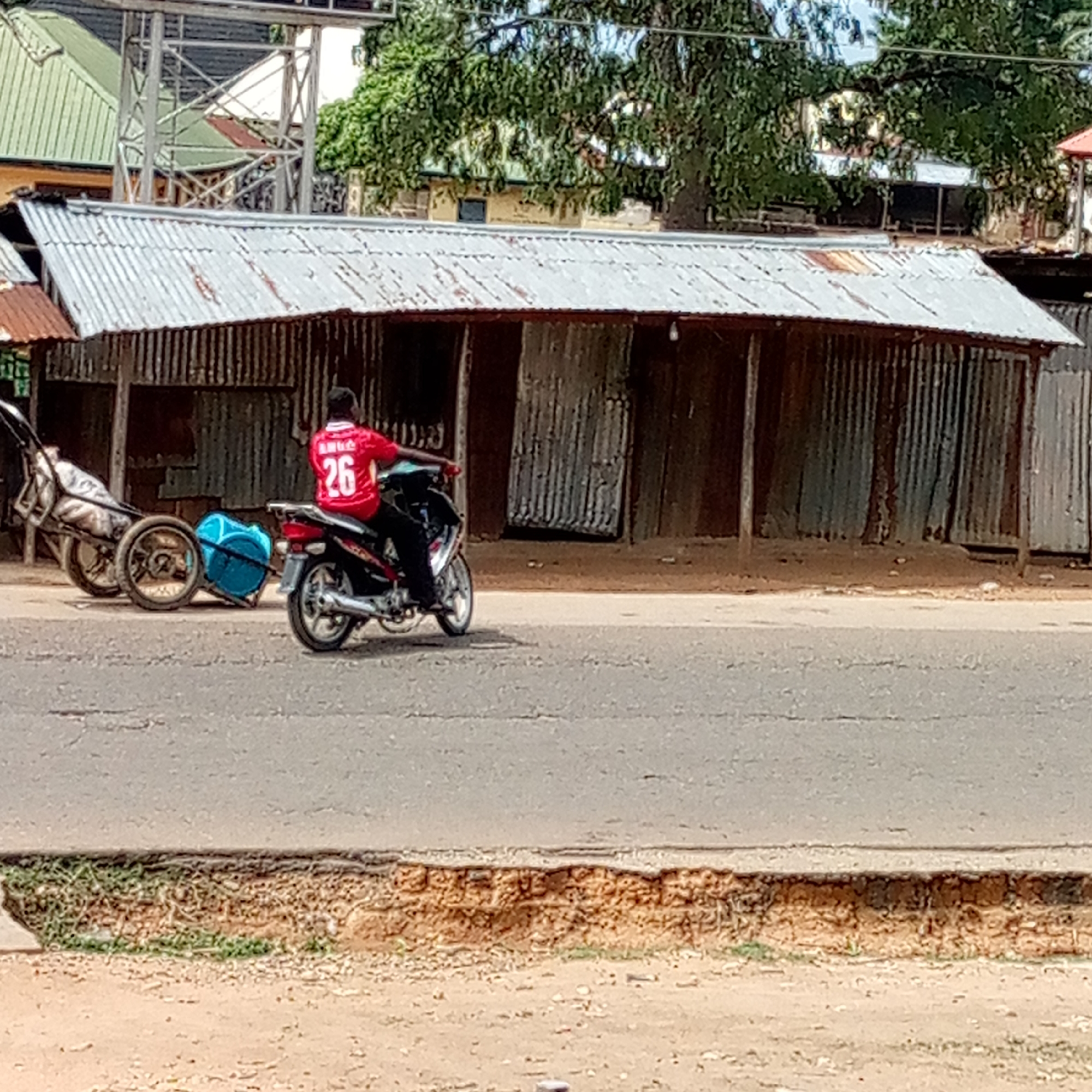 A street scene captures a person riding a motorcycle past corrugated metal structures. In the foreground, a man, viewed from behind, wears a red jersey with the number "26" and some indistinct text (possibly "AMKA") on his back. He is riding a black and red motorcycle, to which a blue plastic container or barrel is attached and towed behind. To the left of the motorcycle, a two-wheeled wooden cart or trailer is parked, containing a white sack or material.
The background features several buildings. Immediately behind the motorcycle and cart is a row of structures constructed from corrugated metal sheets, some appearing rusted and weathered, indicating a makeshift or industrial aesthetic. These buildings have corrugated metal roofs, also showing signs of age and rust. Further in the background, more substantial buildings with green and red roofs are visible, interspersed with lush green trees and dense foliage. A tall metal lattice structure, possibly a power line tower or antenna, is also discernable in the upper left.
The road in the mid-ground is paved asphalt, showing some cracks. Below the road level, there is an exposed dirt bank with visible erosion and some sparse green vegetation, leading down to a lower dirt ground level. The overall impression is a daytime scene in what appears to be a busy, possibly developing, area.