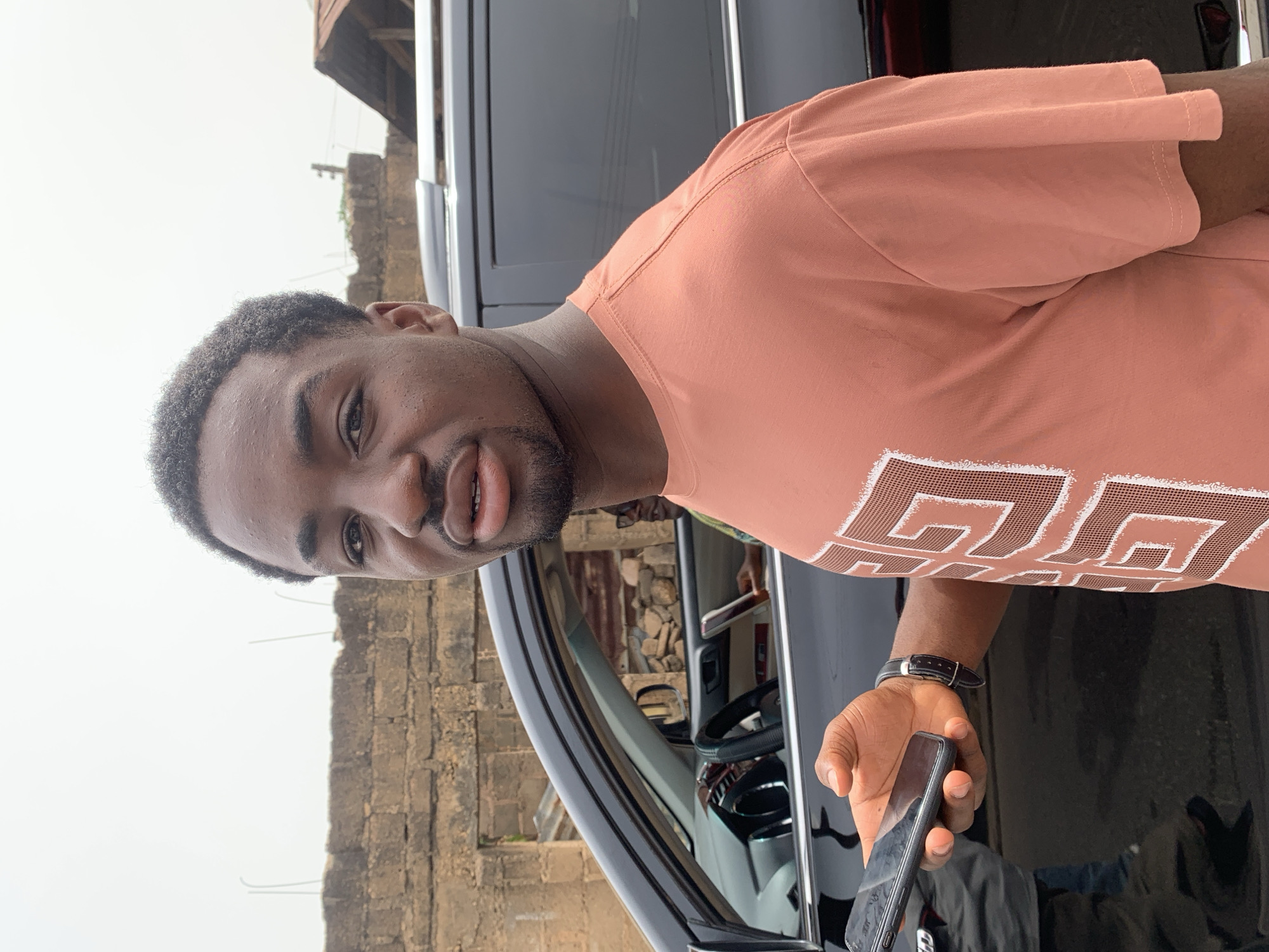 A medium, close-up, rotated shot shows a man in front of a vehicle. The man, who has short black hair, facial hair, and dark skin, is wearing a salmon-colored t-shirt with a geometric design on the front. He is holding a black mobile phone in his right hand. The vehicle is a black SUV. The background includes a weathered brick wall and a partial view of a building. The location is potentially Jos, Nigeria.