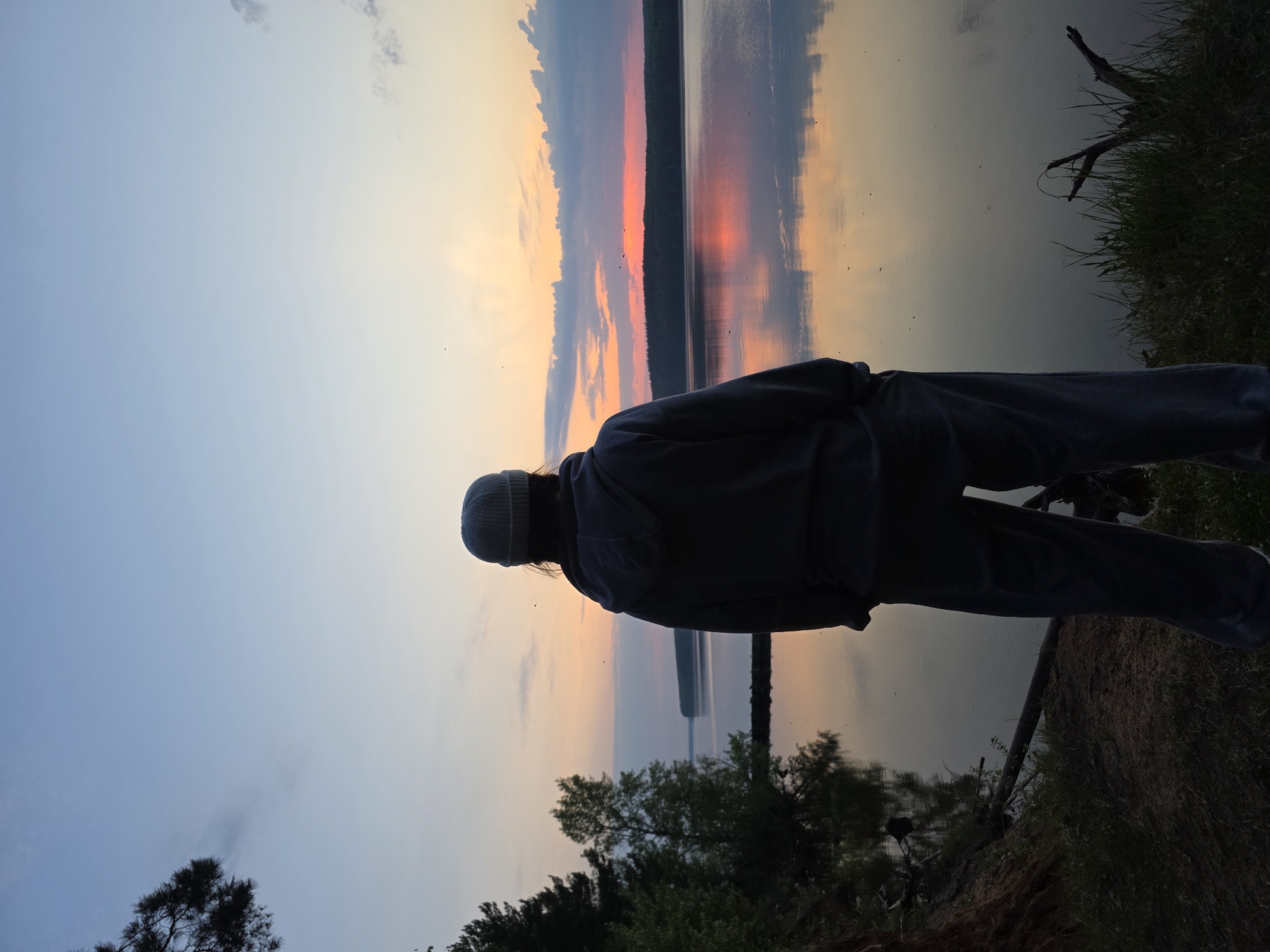 A silhouetted person stands on the bank of a body of water at sunset, the reflection of the sky visible in the water. The location appears to be Chaikovsky, Russia. The person is wearing a knit hat and a jacket, with their back to the camera. To the right, there is grass and a fallen branch. Below, there are trees. The sky is colored with hues of blue, orange, and pink.