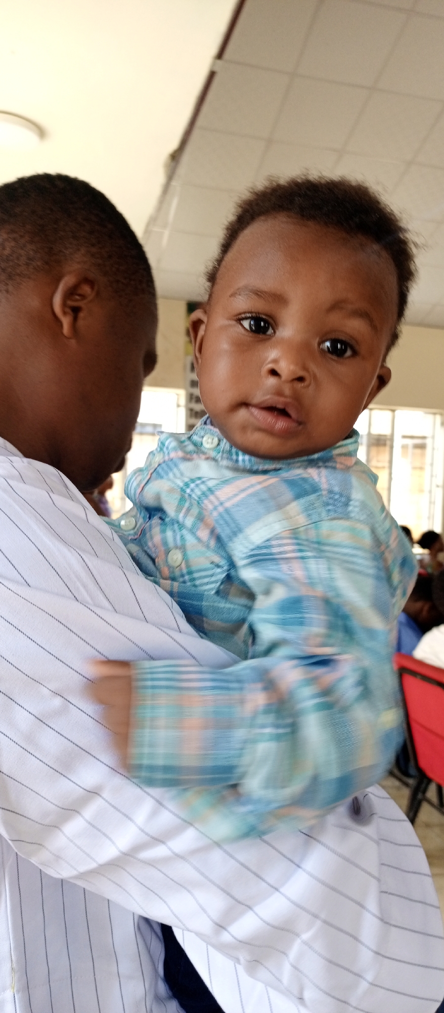 A medium shot captures a dark-skinned man holding a baby in what appears to be an indoor location in Keffi, Nigeria. The man, seen from the back, has a close-cut haircut and wears a white shirt with thin blue stripes. The baby, held facing the camera, is dressed in a blue and orange plaid shirt. The baby has dark eyes and hair. The background includes elements such as a ceiling with light fixtures, windows, red chairs, and other people, suggesting a gathering or public space.