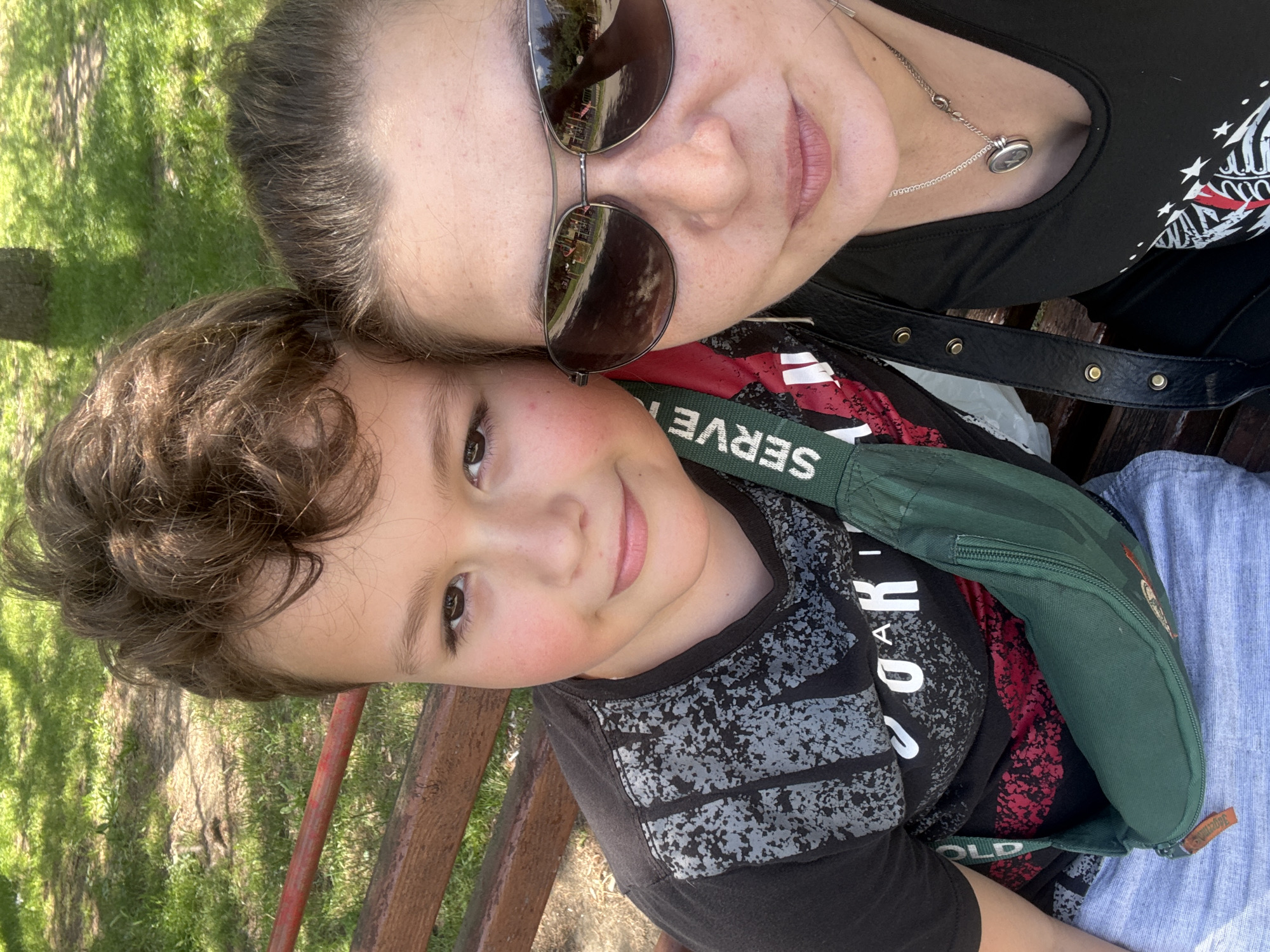 A woman and a young boy are posing for a selfie outdoors, likely sitting on a park bench. The woman is wearing sunglasses, a black top, and a necklace with a circular pendant. The boy, who is resting his head against hers, is wearing a black t-shirt with a speckled design. A green fanny pack with the word "SERVE" is positioned across his body. The background consists of grass and trees, suggesting a park setting. Without further information, it's not possible to confirm the specific location as Varna, Bulgaria.