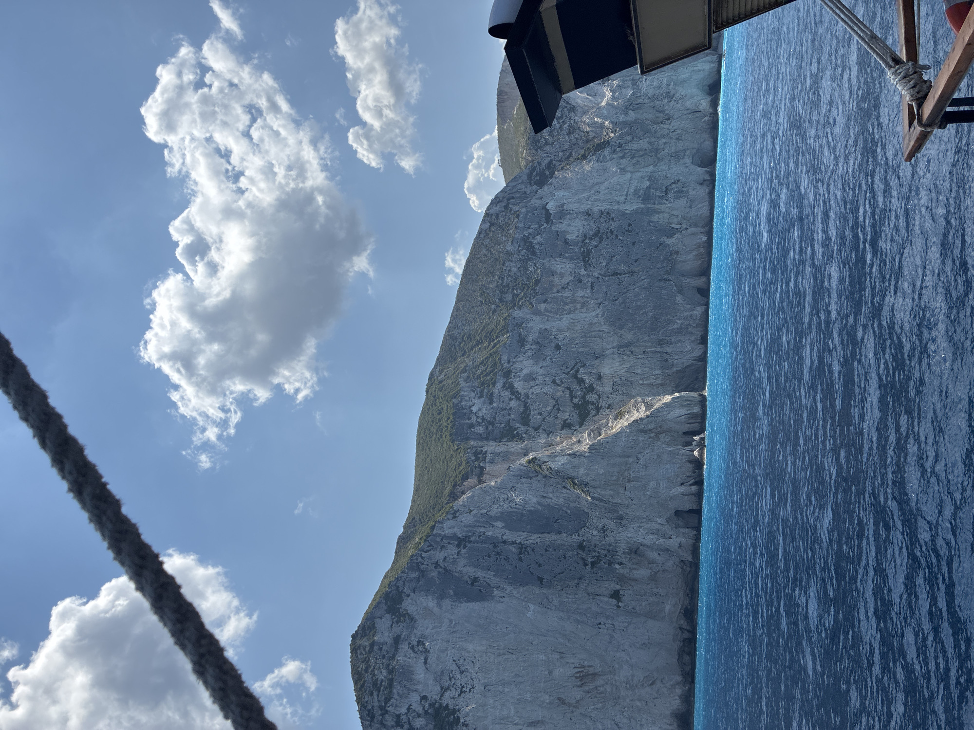 The image captures a view from a boat looking towards a towering, sheer cliff face composed of light-colored rock with patches of green vegetation on its upper slopes. The sea in the foreground is a vibrant, deep blue, with small ripples on its surface. In the upper left of the frame, a thick rope is visible, stretching diagonally across the sky. The sky itself is a clear blue, dotted with fluffy white clouds. Part of the boat's structure, including a dark-colored overhang and wooden railing with rope, is visible on the right side of the image. The scene depicts a coastal landscape, likely in a Mediterranean region, characterized by dramatic cliffs and clear, blue water. The location is the coastline near Elaties Municipal Unit, Greece.