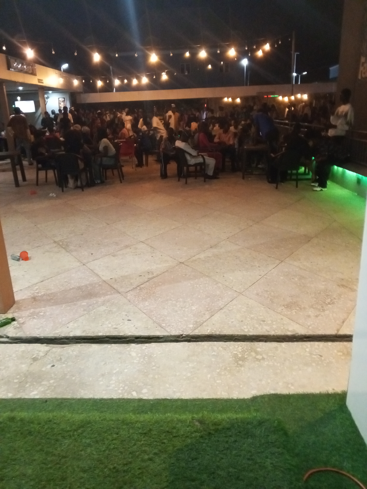 The image depicts an outdoor gathering at night in what appears to be a commercial or social venue. Many people are present, seated at tables and chairs. Overhead, string lights illuminate the scene. In the foreground is tiled flooring with artificial turf along the bottom of the frame. Near the foreground, scattered trash, including a plastic cup and a soda can, is visible. The location is likely Adankolo, Nigeria.