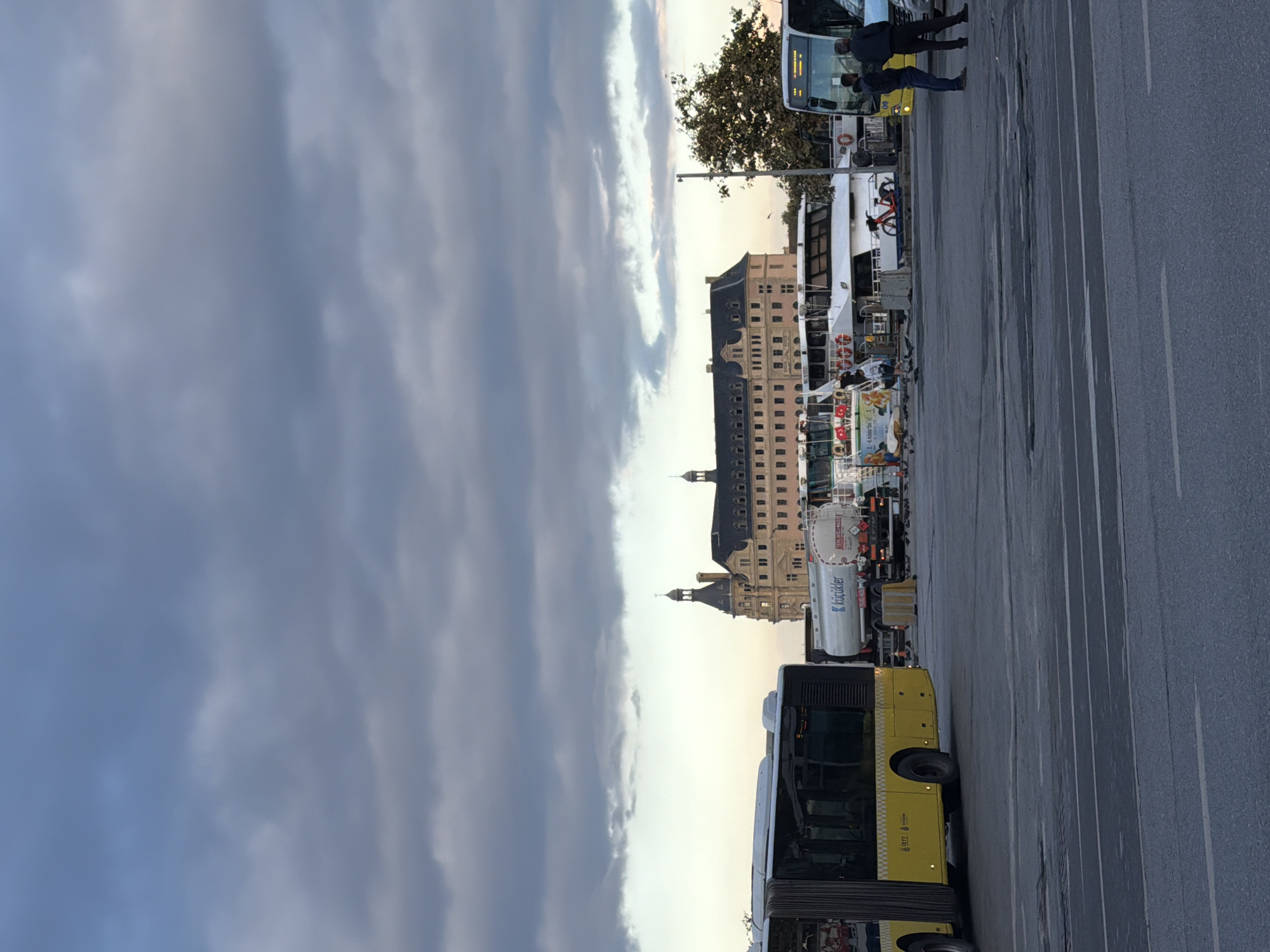The image captures a scene at the Kadıköy ferry terminal in Istanbul, Turkey. The sky is overcast with dramatic clouds. In the foreground, a yellow city bus is parked. Behind it, a large tanker truck is visible, along with several ferries docked. Two individuals are standing near a yellow and white bus on the right side of the frame. The historic Haydarpaşa Train Station building, with its distinctive architecture, forms the backdrop of the scene. The overall atmosphere is that of a busy transport hub under a moody sky.