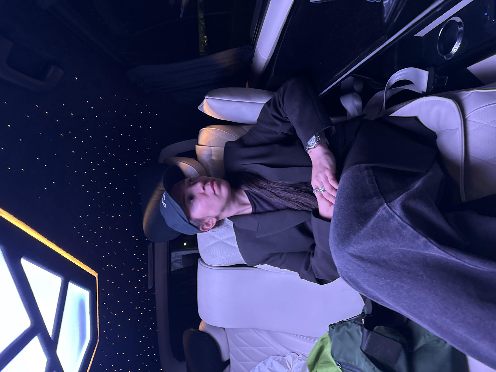 A person wearing a baseball cap, blazer, and casual trousers reclines in a car seat, likely a limousine, based on the interior's design. The car features quilted white leather seats, a dark interior with pinpoint ceiling lights resembling stars, and a modern window design illuminated with blue-white light. The scene is indoors and the person appears relaxed. The location is unlikely to be in Arnavutköy, Turkey, as that is not deducible from the visual information.