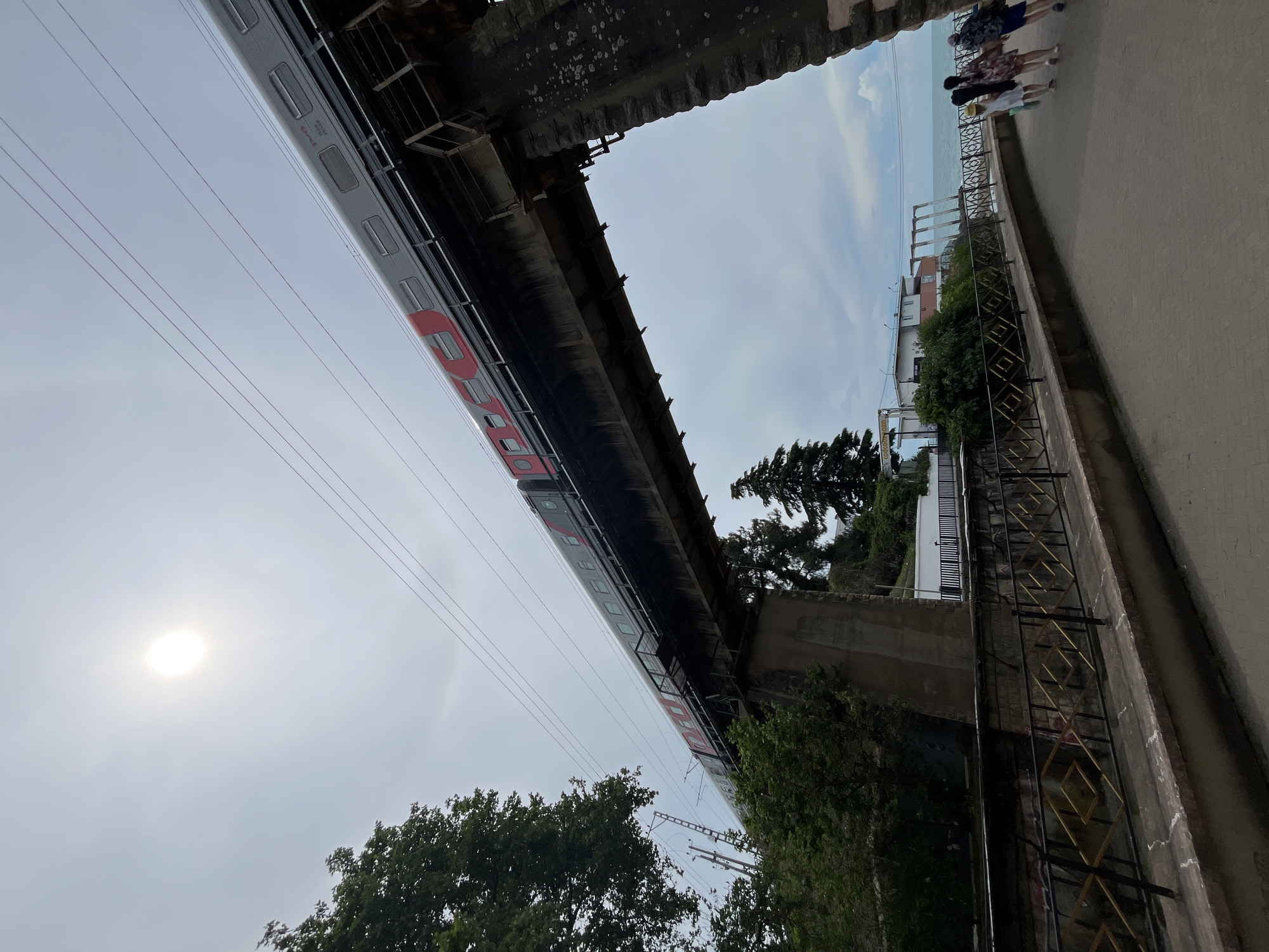 The image shows a train traveling on a viaduct over a pathway next to the sea. The train is silver with red accents and the letters "РЖД" are visible on its side. The viaduct is made of concrete and stone. On the pathway below, several people are walking towards the right of the frame. Some of them are carrying what appear to be musical instrument cases. The sea is visible in the background, with buildings and trees along the coast. The sun is visible in the upper left quadrant of the image, partially obscured by clouds, casting a bright light. The overall scene suggests a scenic coastal location in Sochi, Russia, possibly a promenade or public area near a train line.