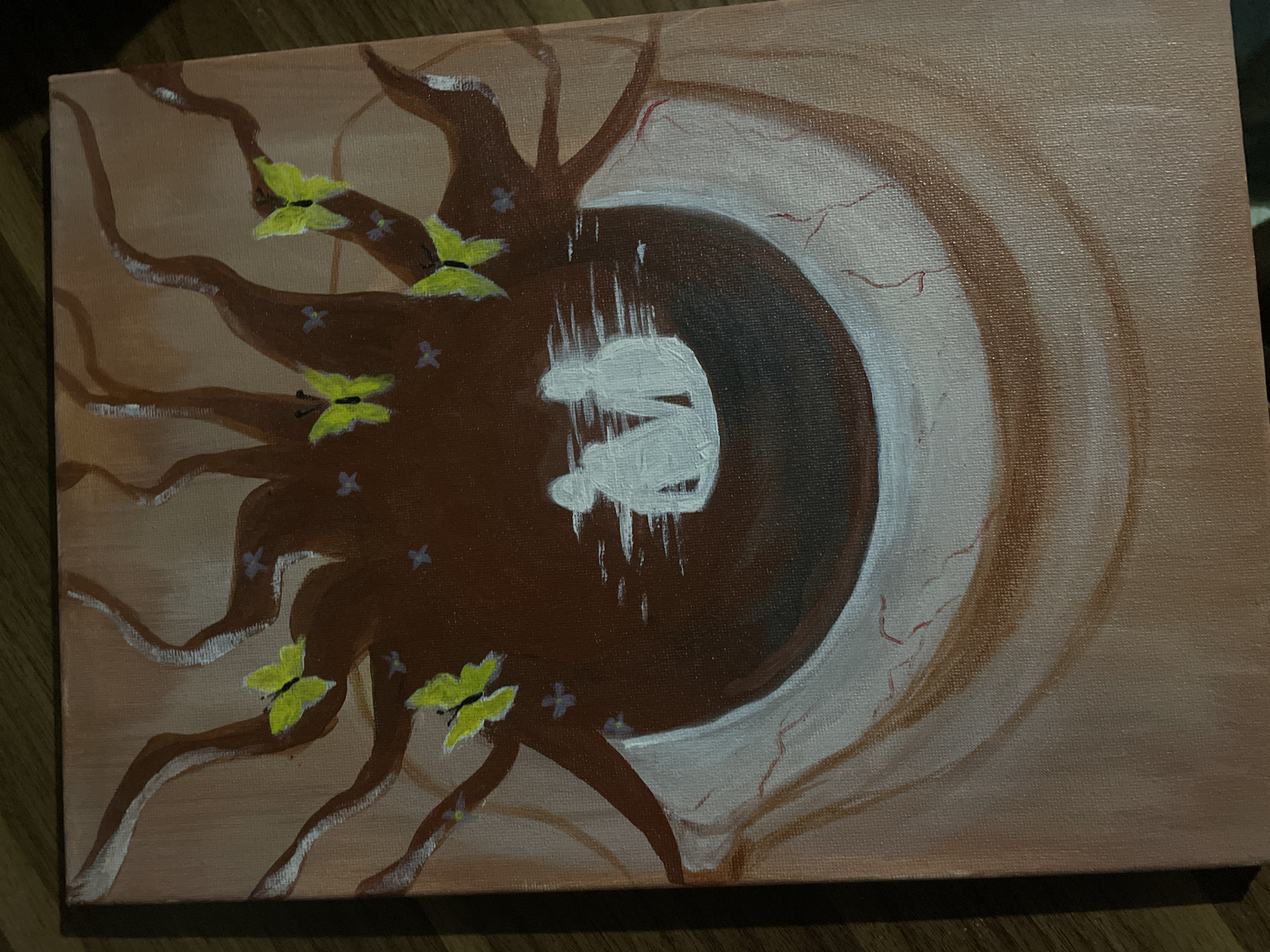 An acrylic painting on canvas depicts a surreal, stylized human eye as its central subject, angled towards the right. The large iris is a deep dark brown, and within its center, a white, abstract figure appears to be falling or emerging, rendered with vertical brushstrokes that suggest motion or dripping. The sclera of the eye is a light, fleshy beige, marked with thin, red, vein-like lines that extend outwards.
Extending from the left side of the canvas and wrapping around the dark brown iris are numerous thick, gnarled, dark brown root-like or branch-like structures. These structures vary in thickness and some have lighter white accents along their edges, suggesting highlights or organic textures. Perched on and around these brown forms are several bright yellow butterflies with black bodies, depicted with a simple, illustrative style. Scattered among the brown roots are small clusters of delicate, light purple-blue, four-petaled flowers or star-like shapes.
The background of the painting, both behind the roots and around the eye, is a soft, sandy beige, subtly transitioning to slightly darker concentric rings towards the right edge of the canvas, which mirrors the circular form of the eye. The painting utilizes an earthy color palette of browns, beiges, and whites, accented by the vivid yellow of the butterflies and the subtle purple of the flowers. No visible text is present in the artwork. The canvas texture is discernible throughout the image. The lighting in the photograph of the artwork is somewhat dim, creating subtle shadows.