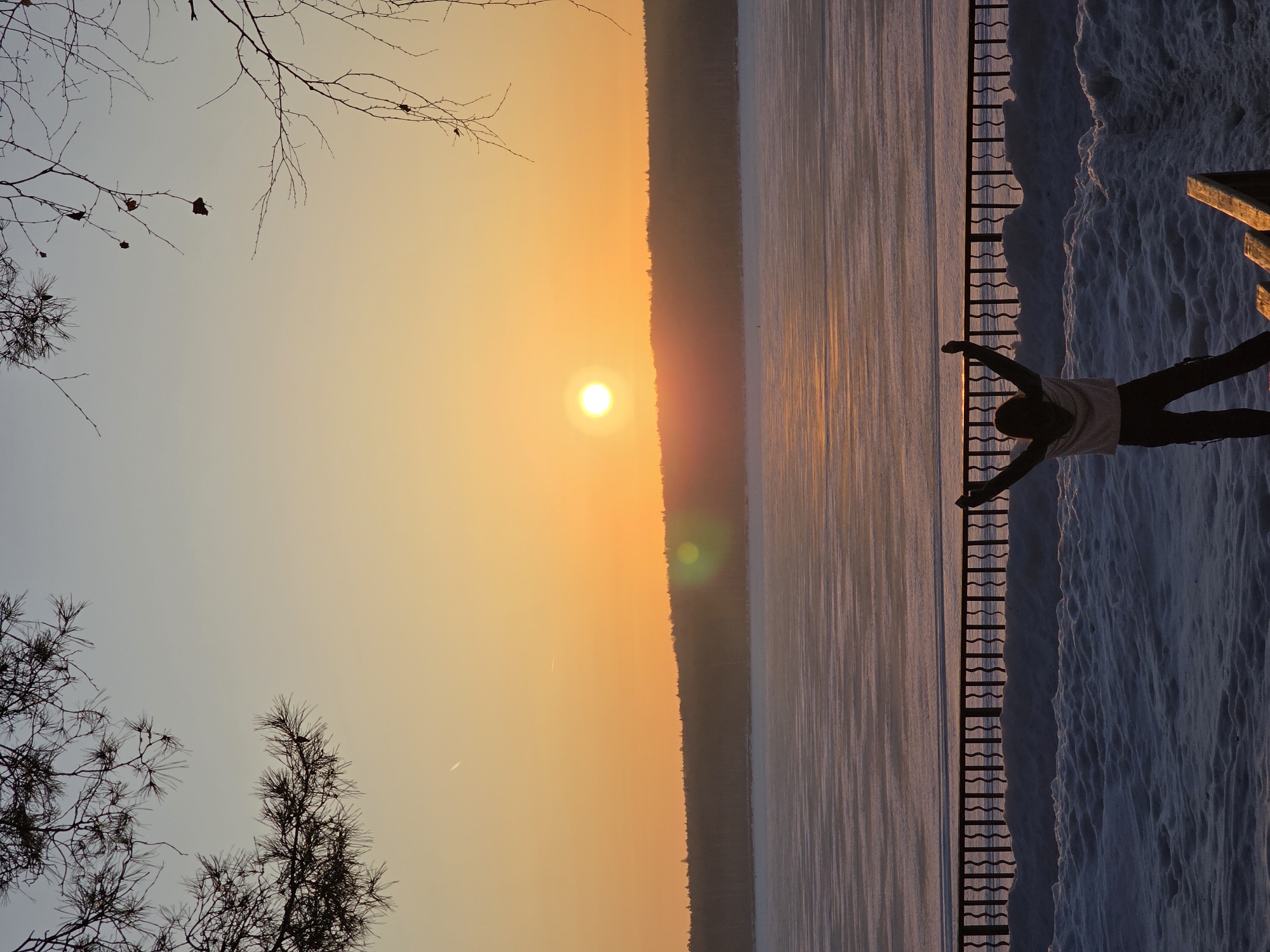 The image depicts a winter sunset over a frozen body of water, likely the Kama Reservoir in Chaikovsky, Russia. The sky exhibits an orange and yellow gradient with a bright, low-hanging sun near the center-left. Two faint white contrails are visible in the upper sky. The horizon is defined by a dark, silhouetted line of forested hills on the far bank. The extensive body of water is covered in textured ice, reflecting the warm hues of the sunset.
In the mid-ground, a dark metal fence with vertical, undulating bars separates the snowy foreground from the frozen water. The foreground is covered in uneven, deep snow. In the lower right corner, the corner of a wooden structure, possibly a bench or steps, is partially visible.
A single individual, silhouetted against the bright sunset, stands in the foreground, facing away from the viewer towards the frozen water and sunset. The person is dressed in dark pants and a light-colored top, with both arms raised above their head in a widespread gesture. Silhouetted bare tree branches are visible in the upper right, and pine branches with needles are present in the upper left corner of the frame.