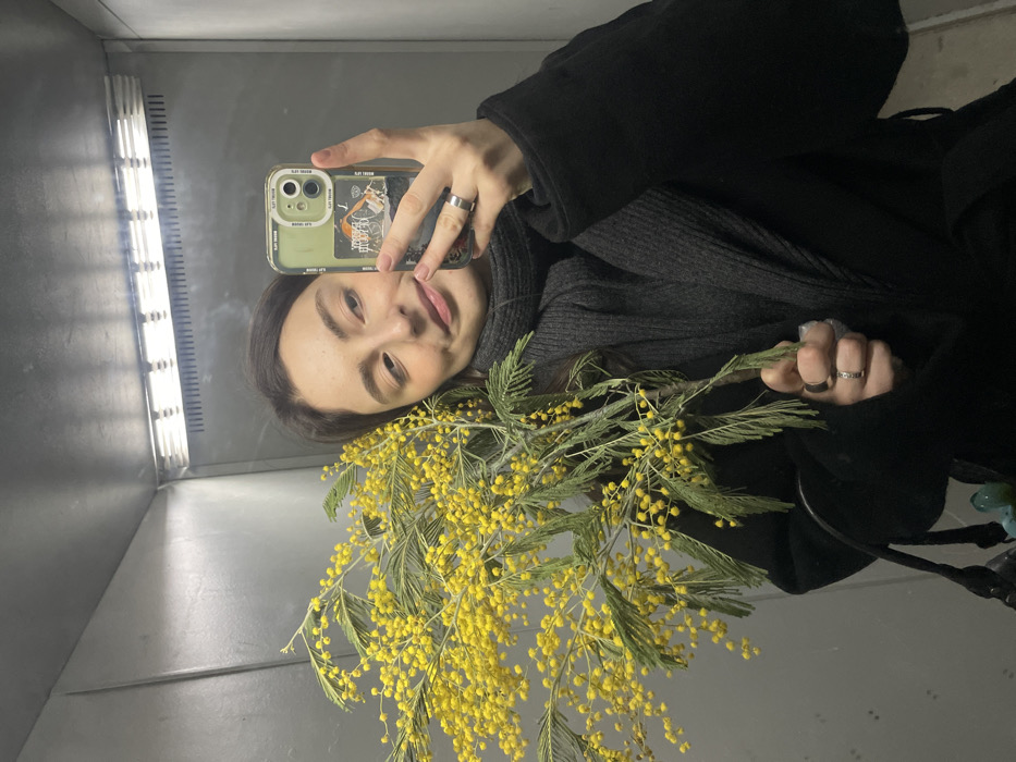 A female subject is depicted taking a mirror selfie within a metallic elevator in Moscow, Russia. She is centrally framed, looking slightly past the camera lens with a subtle smile. The subject has dark hair and fair skin, dressed in a dark coat and a grey ribbed scarf or turtleneck. In her left hand, she holds a large bundle of yellow mimosa flowers with small, spherical blossoms and green feathery leaves. Her right hand operates a smartphone in a translucent green case, which displays multiple stickers or cards, one featuring a stylized "M" logo, indicative of the Moscow Metro system. A silver ring adorns her right ring finger. The elevator interior consists of reflective brushed metal panels on the walls and ceiling, illuminated by a horizontal fluorescent light fixture. A section of a dark bag with small blue-green decorative elements is visible in the lower left foreground. The exact Moscow Metro station is not identifiable from the provided visual information.