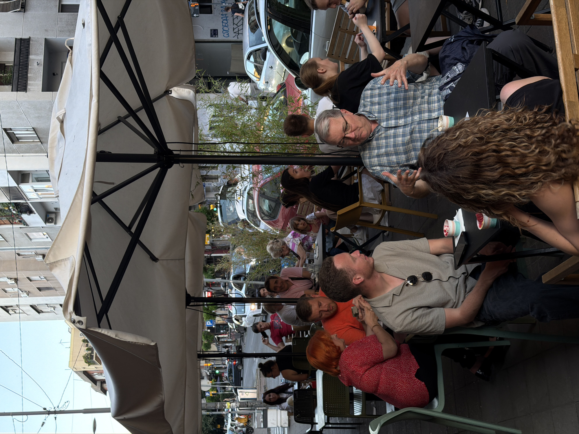 The scene depicts an outdoor cafe setting in the Vracar Urban Municipality, Serbia. Several groups of people are seated at tables under large umbrellas. The perspective shows a street with parked cars and buildings lining the sides.
People present in the scene include a man wearing glasses and a blue plaid shirt, conversing with a woman with curly hair, a man wearing a beige shirt and jeans, and a woman in a red polka dot dress. Other people are seen further down the street, some standing or walking.
Objects present include tables, chairs, umbrellas, buildings, parked cars, and assorted cafe accessories. The actions occurring are people talking, sitting, and interacting. The environment appears to be a typical urban street with cafe seating extending onto the sidewalk.