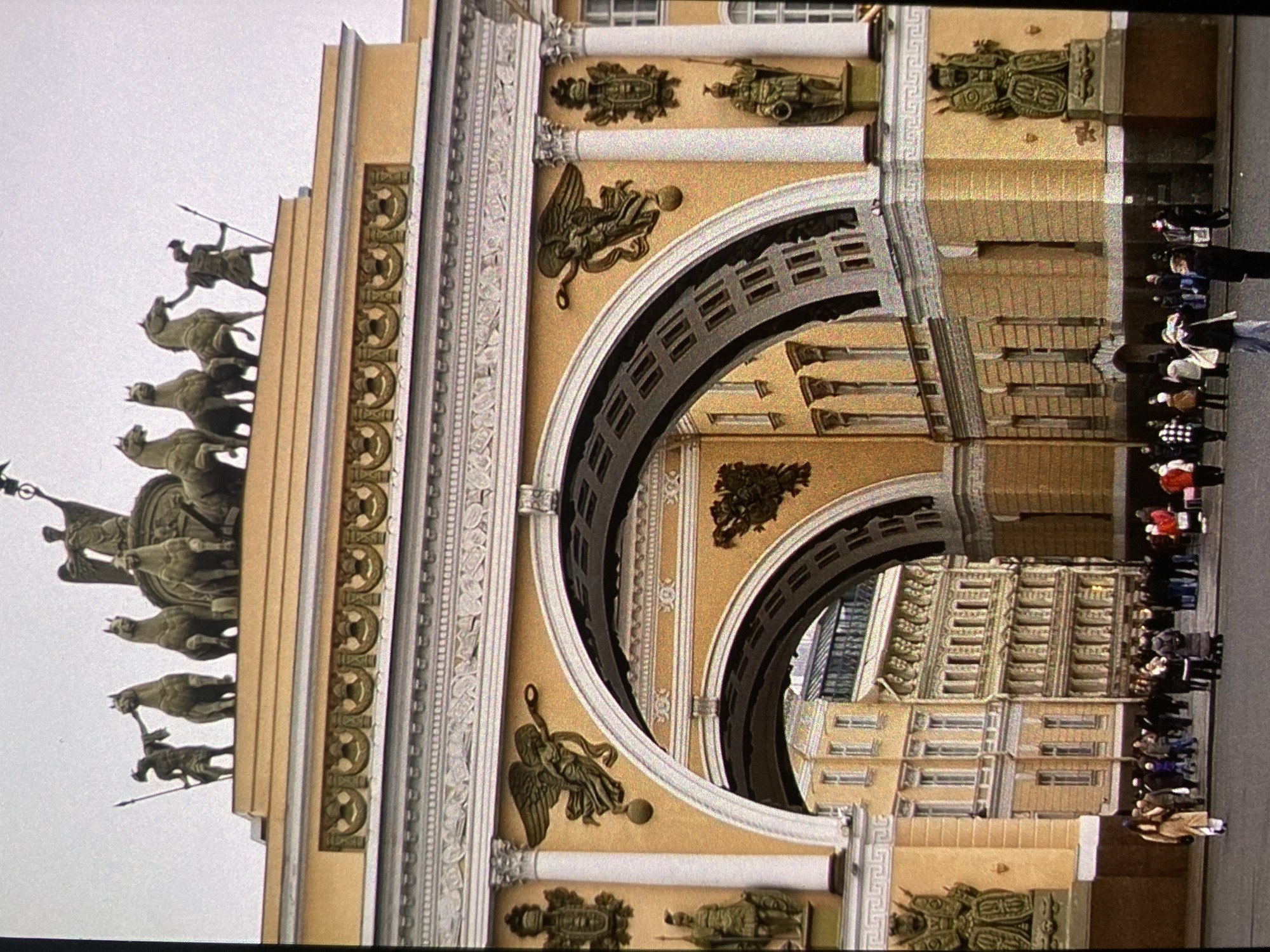 The image displays a close-up, angled view of the Triumphal Arch of the General Staff Building in Saint Petersburg, Russia. The grand Neoclassical structure is characterized by its pale ochre facade and rich white and golden architectural ornamentation.
The most prominent feature is the bronze quadriga atop the arch, depicting a chariot drawn by six horses in dynamic motion. A figure, likely a Roman goddess of Victory, stands in the chariot, holding a spear or staff. Below the main cornice and around the archways, the building is adorned with elaborate sculptures and bas-reliefs. These include winged figures (possibly Nikes) holding various emblems, as well as detailed military trophies and weaponry, such as shields, helmets, and cannons, set into the facade. The archways themselves are massive, with a larger outer arch encompassing a slightly recessed inner arch. Ornate columns and pilasters with intricate capitals are visible, adding to the grandeur.
Through the arch, parts of another building with numerous windows and decorative elements can be seen, hinting at the vastness of Palace Square beyond. On the right side of the image, a group of people, likely tourists, are gathered on the ground level, some appearing to stand in a line. They are dressed in coats and jackets, suggesting a cool or temperate climate. The sky is overcast and bright, indicating it is daytime. No discernible text is visible on the building or surrounding elements. The image is presented rotated 90 degrees counter-clockwise.