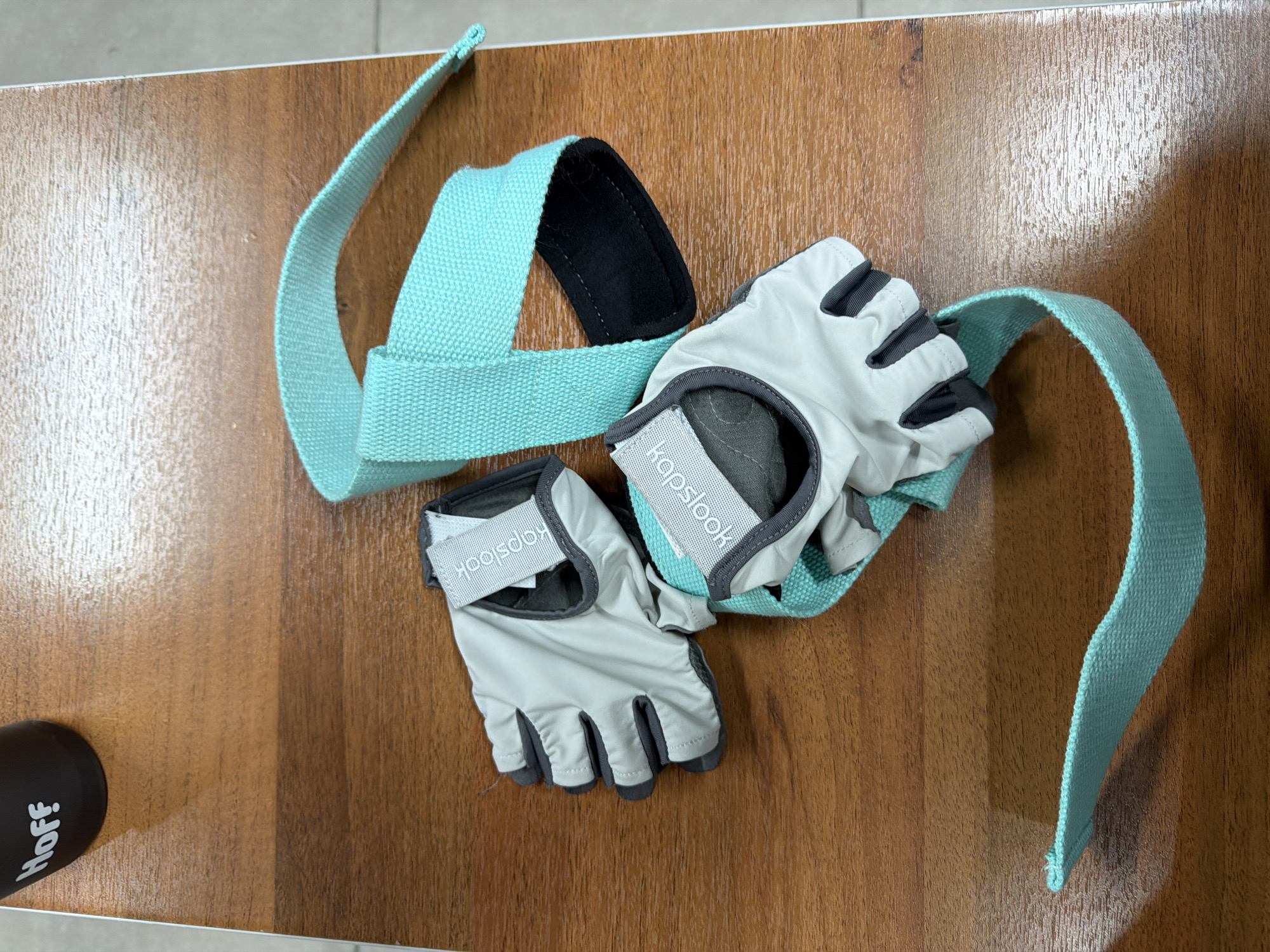 The image shows a pair of light gray weightlifting gloves with dark gray accents and a light blue weightlifting belt on a brown wooden surface. The gloves are fingerless, and the brand name "Kapstool" is visible on the wrist strap. The weightlifting belt is a loop with a black fabric pad on one section. Part of a black water bottle with the name "Hoff" in white lettering is visible in the lower left corner of the image. The scene is indoors. There is no indication of the location being Kazan, Russia.