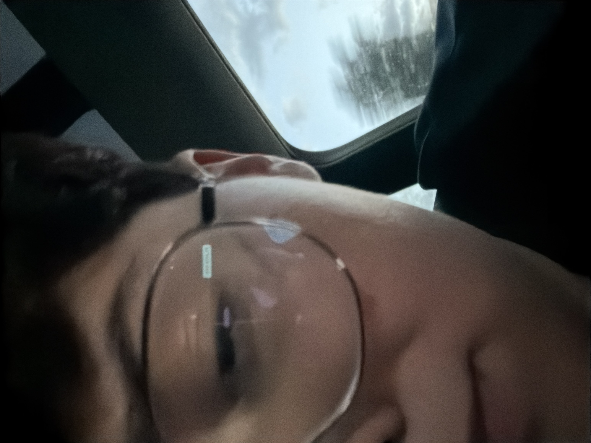 The image shows a close-up, slightly distorted, side-profile view of a person wearing large, round-framed glasses. The person's left eye is visible through the lens, and a portion of their face, including their cheek and mouth area, is also visible. A faint reflection or sticker is present on the left lens of the glasses. The person appears to be inside a vehicle, indicated by the car interior visible in the background, specifically the A-pillar and the window area. The window shows a blurred view of trees and a cloudy sky, suggesting the vehicle is in motion. The image quality is grainy. There is no information within the image to indicate the location is the city of Moss, Norway.
