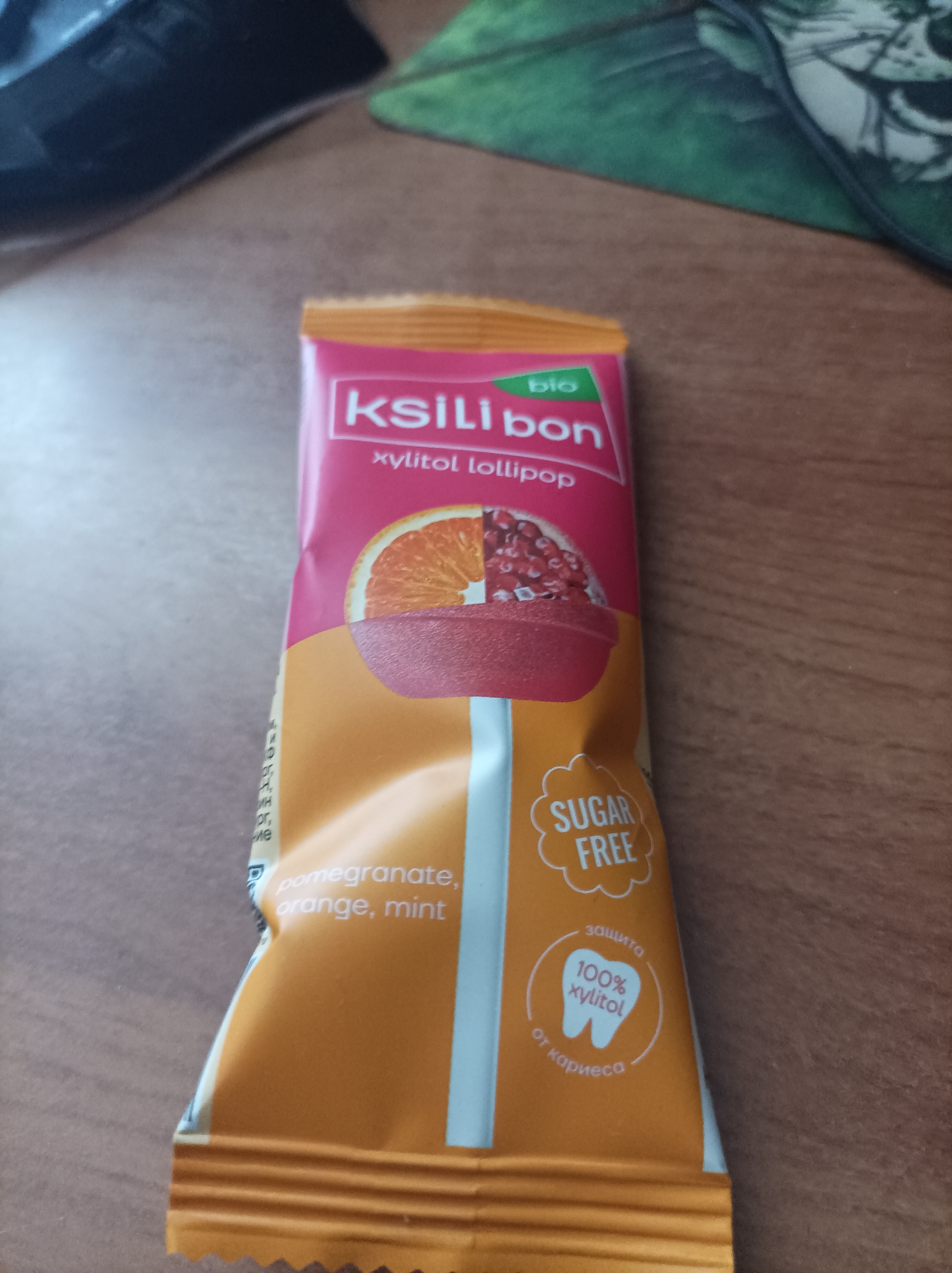 A single KSilbon xylitol lollipop package is centered and slightly angled on a light brown wooden surface. The package, primarily in shades of pink and orange, features a white horizontal stripe across its middle. The top pink section displays the brand name "KSilibon" in white capital letters, with a small green "bio" label above the 'i'. Below the brand, "xylitol lollipop" is written in smaller white text. An illustration of a sliced orange half and pomegranate seeds is depicted on a red lollipop stick within this pink section. The lower orange section of the package lists the flavors: "pomegranate, orange, mint" in white text. A white cloud-shaped emblem in the lower right corner states "SUGAR FREE," while a circular white emblem beneath it features a tooth icon and the text "защита 100% xylitol от кариеса" (protection 100% xylitol from cavities in Russian). The package exhibits minor creasing. In the out-of-focus background, a dark, indeterminate object is visible in the upper left, and a green patterned object, possibly a mouse pad, is partially visible in the upper right.
