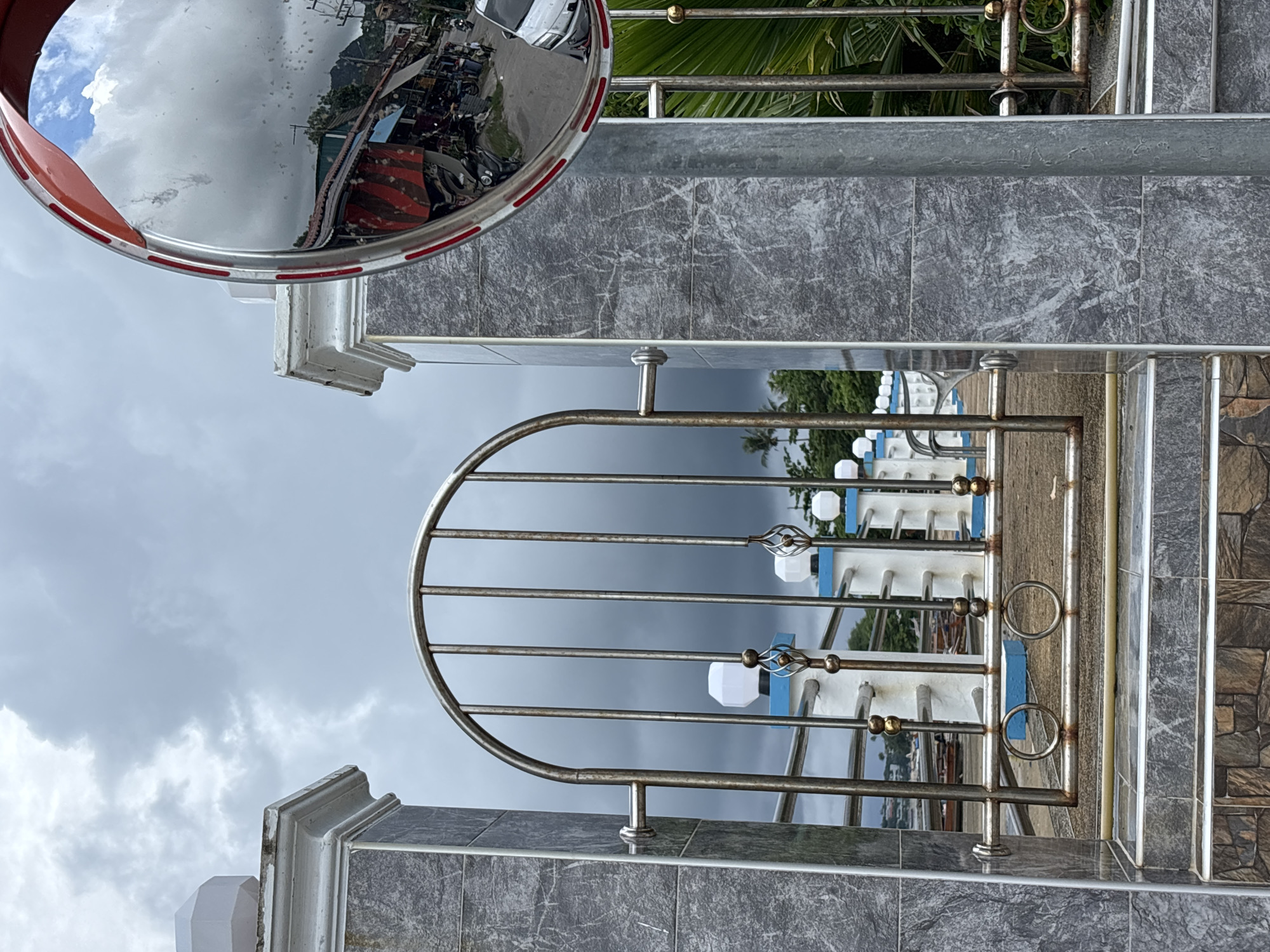The image depicts an outdoor scene with a focus on a metal gate and decorative pillars, likely situated in Rawai, Thailand, given the context of the surrounding environment. The gate, made of polished stainless steel, features vertical bars and an arched top, with decorative elements and a handle. It is flanked by two pillars clad in grey, marble-like tiles, topped with white, octagonal structures. Behind the gate, a white railing with blue accents and small, white, hexagonal lights can be seen. The background shows a body of water with several moored boats, suggesting a waterfront or pier location. The sky is overcast with heavy, dark clouds, indicating a possibility of rain. A large, convex traffic mirror, framed in red and silver, is visible on the right side of the image, reflecting a street scene with buildings, parked motorcycles, and a white car. Lush green palm fronds are visible on the far right, adding to the tropical ambiance. The ground in front of the gate is paved with stone tiles. No people are directly visible in the main focus of the image, but the reflection in the mirror suggests the presence of human activity in the depicted street.
