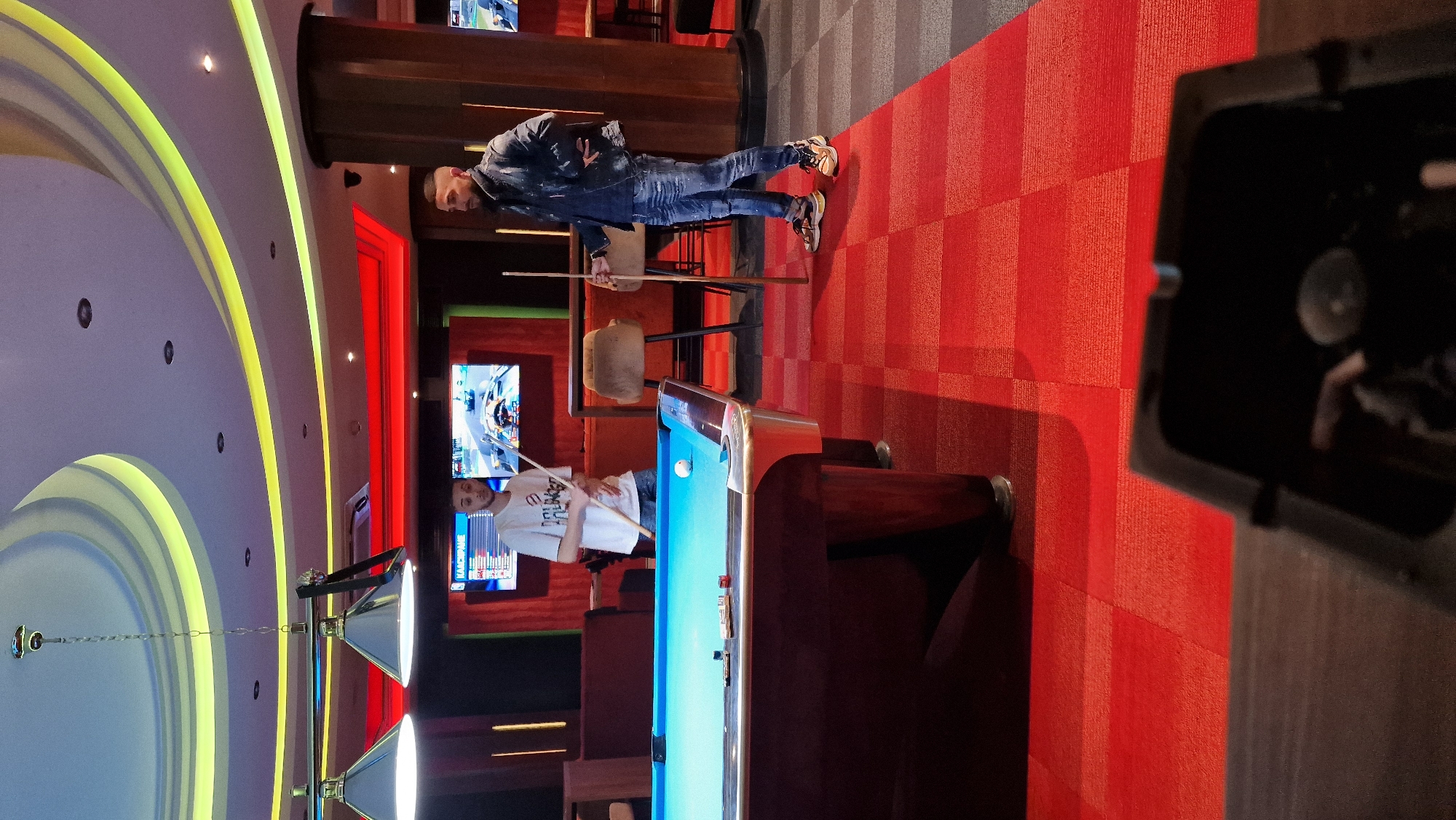 The image shows the interior of a recreational establishment, likely a billiards hall, possibly located in Sofia, Bulgaria. Two men are present. One man, dressed in a denim jacket and jeans, stands near a bar area, holding a cue stick. Another man in a white T-shirt prepares to strike a cue ball on a blue felt pool table. A set of pool table lights hangs above the table. A red-patterned carpet covers the floor. A television screen is visible in the background showing some sort of race.